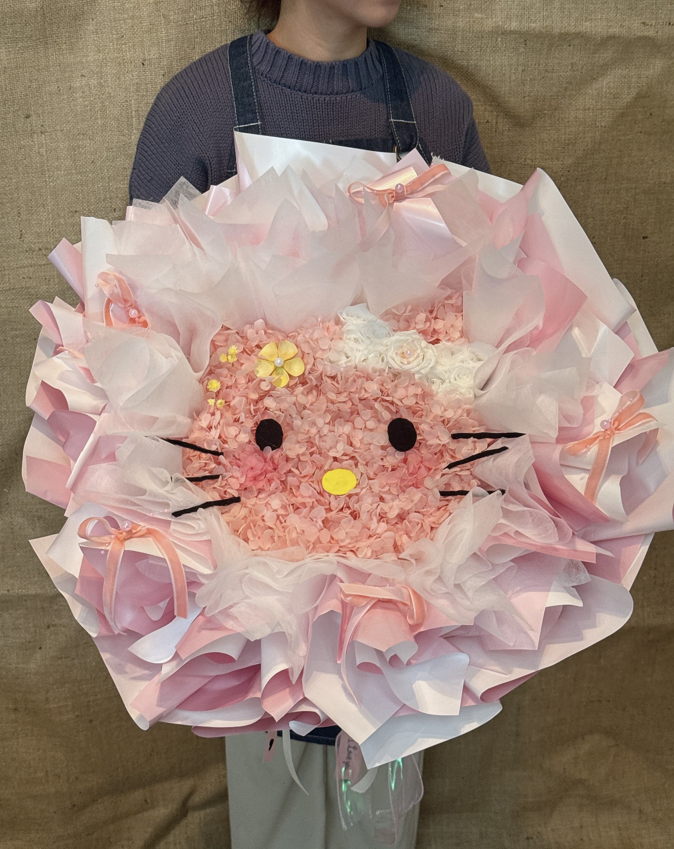 Pink Hello Kitty Bouquet - This bouquet is based on the theme of the popular cartoon character Hello Kitty, carefully made into Hello Kitty's face shape and beautifully packaged. Flower material: The main body of the bouquet is composed of a large number of pink and a little white flowers, which make up the facial outline, bow, ears and nose of Hello Kitty. Usually, full of star flower material is used for long-term preservation. Packaging: Wrapped with multiple layers of white and light pink transparent tulle or organza, creating a fluffy and dreamy Korean bouquet style. Decoration: There is a ribbon at the bottom and may contain a decorative balloon. Meaning: Hello Kitty bouquet is a symbol of girl's heart and love, which is very suitable for expressing love, surprise and special care. The pink tone further strengthens the meaning of romance, gentleness and first love.