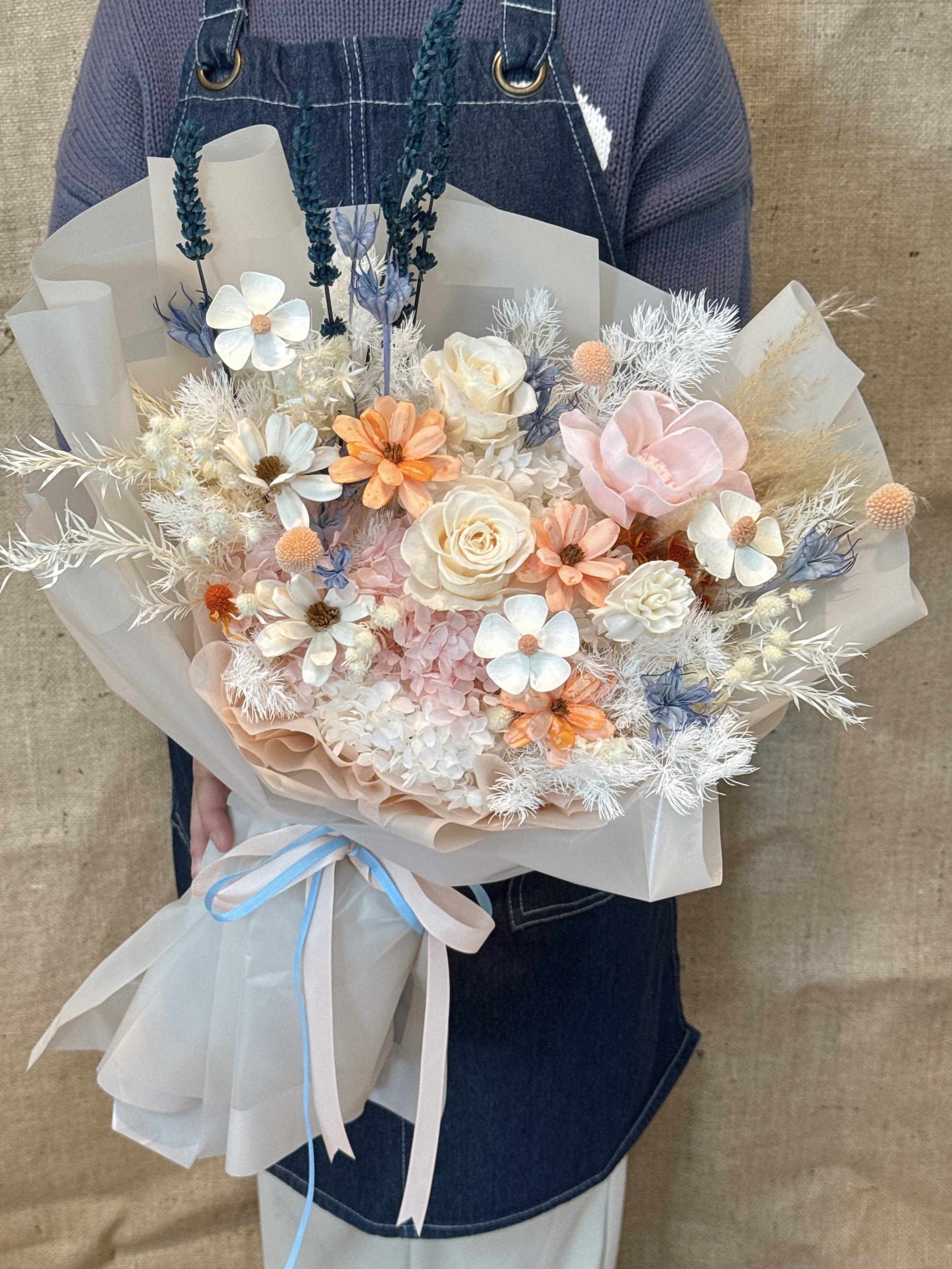 Friendly Bouquet - This is a fresh and natural mix and match bouquet. This bouquet adopts a combination of earthy colors and soft colors, giving people a warm, comfortable and artistic feeling. Flower materials and color matching The bouquet is mainly in soft orange, white, blue and beige: Main tone: soft beige, warm orange and light blue, the color is naturally harmonious. Main flowers: including beige and warm orange roses and small white daisies Decoration: Unique dried flower materials are used, such as blue feather-like or grassy plants, and natural green eucalyptus leaves, adding a sense of hierarchy and bohemian style. Style and packaging Style: Adopt the current popular Korean ins style design, pursuing natural, casual and exquisite beauty. Packaging: It is wrapped with multiple layers of light transparent organza or tulle, which makes the bouquet look fluffy, soft and has a dreamy atmosphere. Decoration: There are light blue and beige ribbons at the bottom, which adds a sense of exquisiteness. Meaning and use Meaning: This bouquet is warm and natural, implying sunshine, hope, comfort and unique love. Occasion: Suitable for birthday gifts, housewarming gifts, holiday surprises or any occasion that need to add a warm and artistic atmosphere.