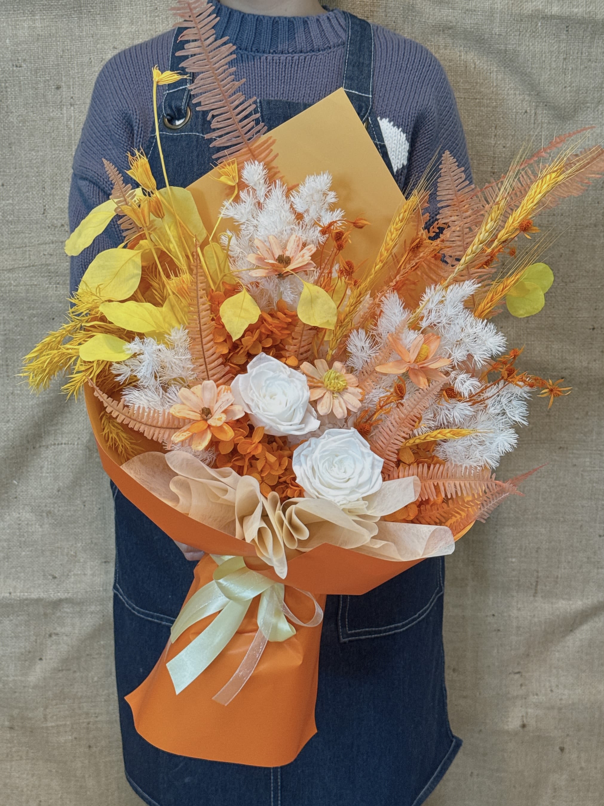 Cheerful Charm - This is an orange-yellow mixed bouquet of immortal flowers/dried flowers, with a warm, bright and energetic style. Composition of flower materials Main flower: mainly orange and yellow immortal roses or similar focal flowers. Filling flowers: including white stars, immortal hydrangeas and small flowers similar to daisies. Texture and lines: A variety of dried wheat ears, oat grass and ferns, as well as fluffy rabbittail grass and reeds, are used to add rich textures and layers. Packaging style The bouquet is wrapped in orange and yellow Korean wrapping paper. The tone is bright and warm, highlighting the sunshine temperament of the flower itself. Finally, it is tied with light beige or white ribbons and decorated with small beads to add a sense of exquisiteness. Symbolic meaning Orange and yellow usually symbolize enthusiasm, joy, vitality, success and friendship. This warm tone combination can boost the spirit and bring positive mood. Due to the use of eternal flowers, this bouquet also represents eternal friendship or lasting enthusiasm. It is very suitable for celebrating achievements, birthdays, giving to friends or any occasion that requires vitality and warmth.