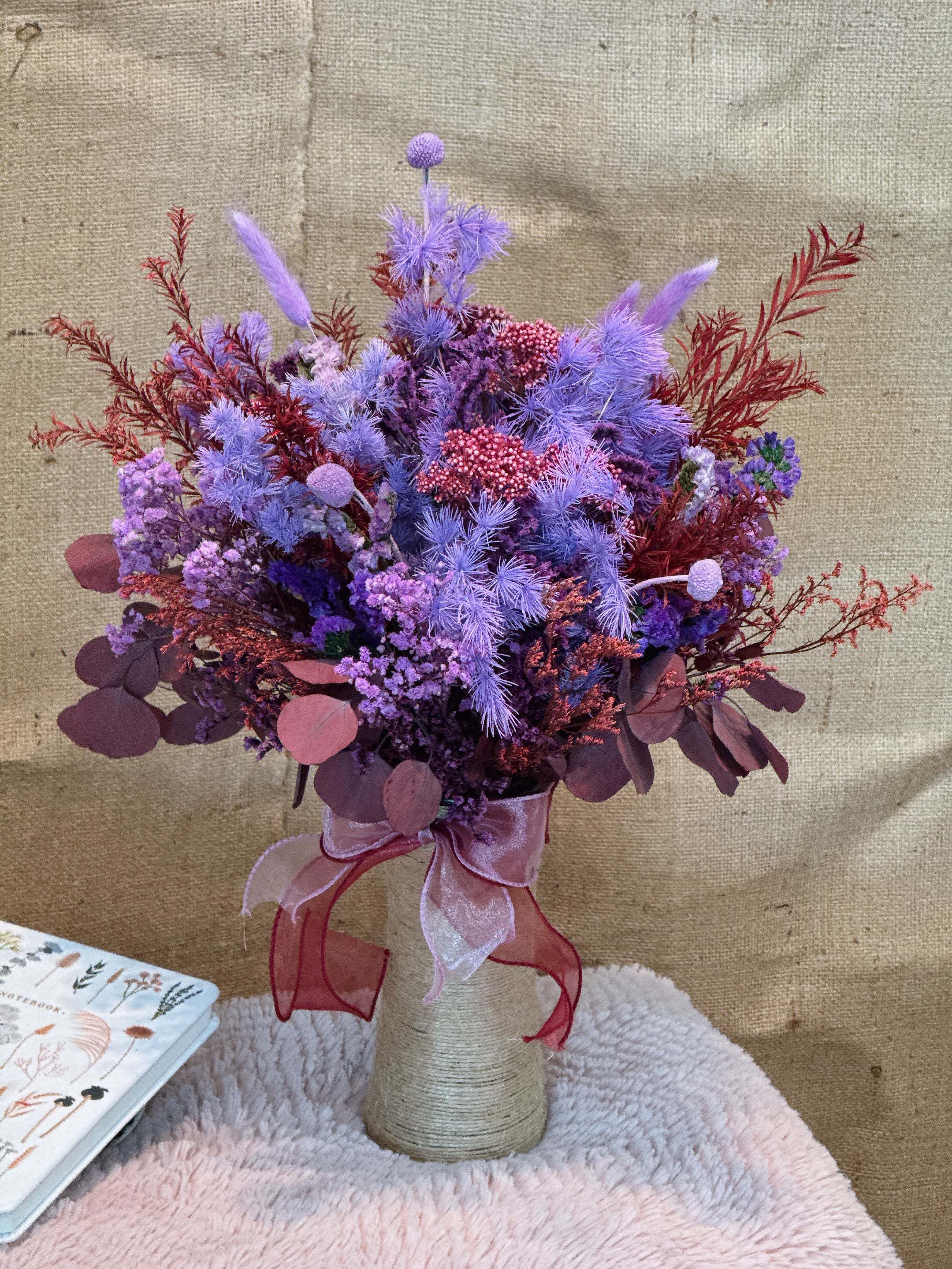 Evening Breeze - This vase is mixed with purple and red dried flowers and flowers, creating a rich sense of hierarchy. The bouquet includes purple lavender-like feather-like flowers, purple cornflowers, blue chrysanthemums, red small fruit flowers and crimson leaves. The vase is wrapped in burlap, and the bottleneck is tied with a pink ribbon bow, adding a warm and elegant atmosphere. The overall style is retro and romantic, which is suitable for decoration or as a gift.