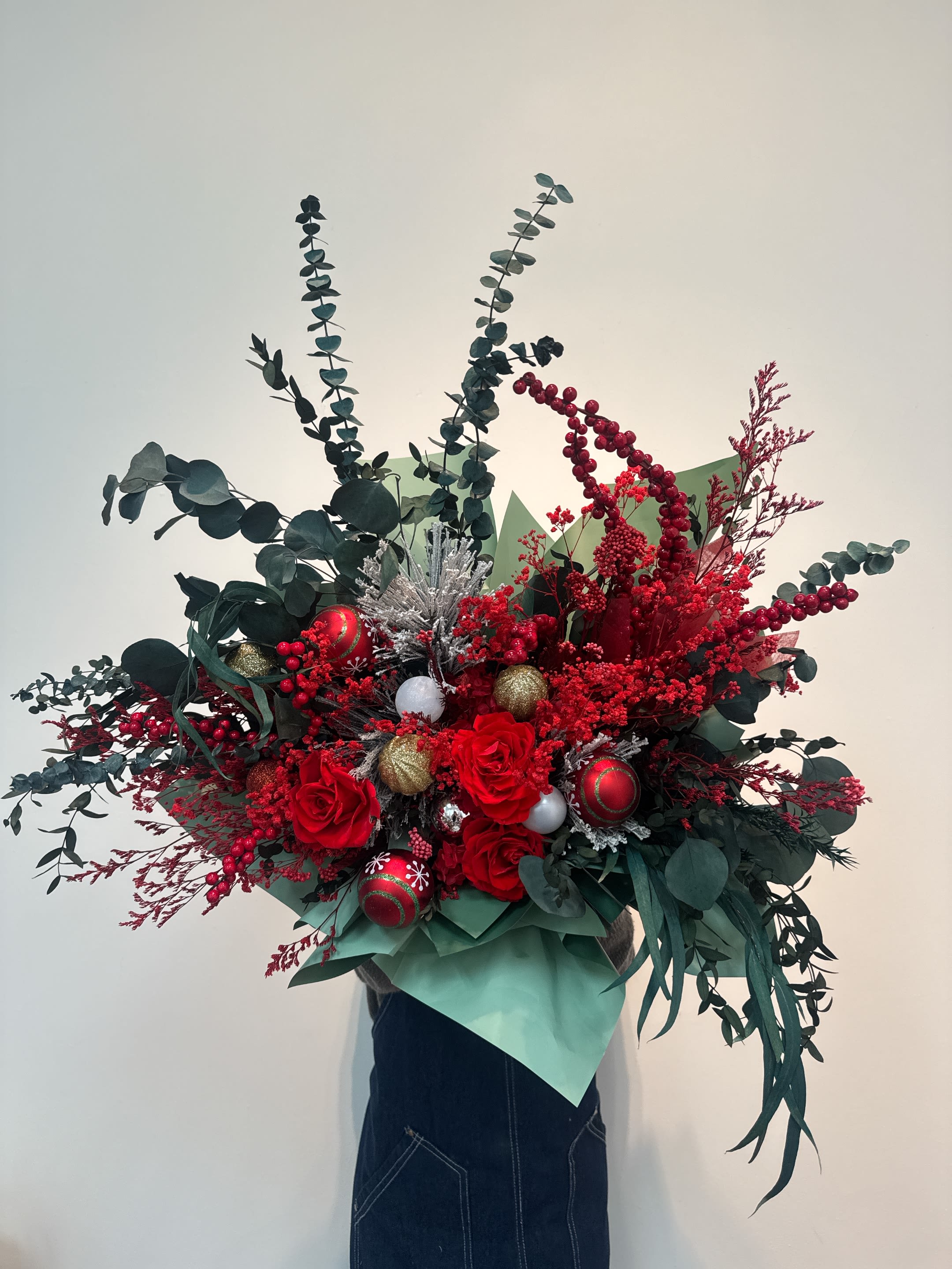 Christmas Arrangement  - This is a Christmas-themed basket, mainly composed of red roses, eucalyptus leaves, red berries and other flower materials. Red roses symbolize love and romance. Eucalyptus leaves (eucalyptus leaves) add a winter atmosphere and a sense of luxury to the bouquet with their unique aroma and elegant silver-green tone. Red berries (such as holly or similar artificial berries) are dotted, creating a strong festive atmosphere. The bouquet also contains silver decorations (such as balls or glittering twigs) and green wrapping paper, which further enhances the festive color of Christmas (red, green and white/silver are the traditional colors of Christmas). This style of bouquet is often used for church Christmas decorations, holiday gifts or company openings.