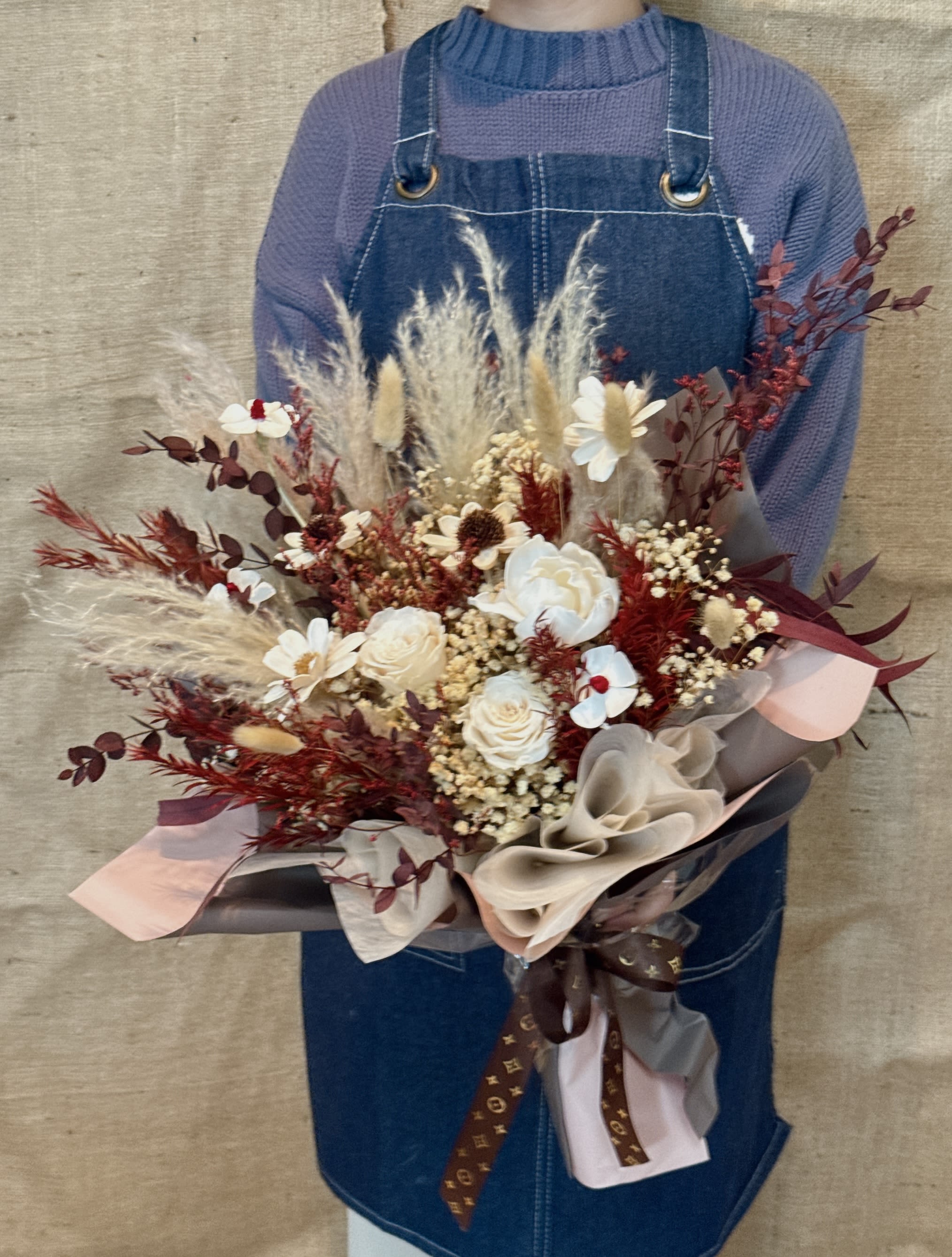 Warm Meadow Bouquet - This is a mixed bouquet of eternal flowers/dried flowers in cream white, crimson and brown tones, with a warm, retro and fashionable sense of bohemian or "Maillard style". Focus flower: creamy white, light beige and dark red immortal roses. Matching flowers and leaves: use white full stars, red dried flowers and dark green eucalyptus leaves as filling. Texture and lines: A variety of naturally dried plants are used, such as dark brown fern leaves, fluffy beige rabbit tail grass, oat grass and pampas grass, which add rich textures and layers. Packaging style The bouquet adopts Korean-style wrapping technique, using multi-layer white or light-colored waterproof wrapping paper as the lining, and the outer layer is matched with brown or khaki cowhide paper packaging. This combination is low-key and warm. Symbolic meaning The cream tone of this bouquet symbolizes purity, gentleness and a new beginning, while the red tone brings enthusiasm and vitality, and the brown tone brings the meaning of warmth, stability and gratitude. Because of the use of eternal flowers, it also represents eternal love or friendship. It is very suitable as a graduation gift, a thank you to your teacher, an anniversary or any occasion that needs to express sincere wishes.