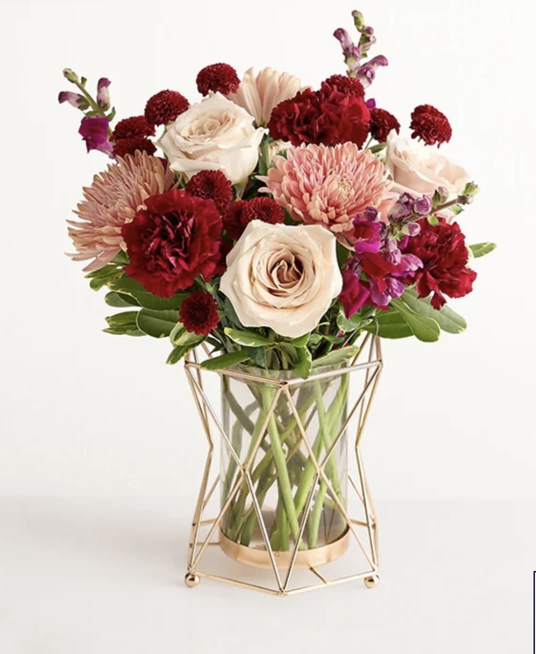 Burgundy Bliss  -  Burgundy Bliss™ Bouquet NEW Autumn’s opulent palette inspired this mixed bouquet, which features pale sandy roses, deep burgundy snapdragons, and more. Adding to the sense of luxury is our choice of vase, whose geometric silhouette is outlined in gleaming gold. More details Product Details: All-around arrangement with sand-color roses, burgundy snapdragons and carnations, peachy pink mum disbuds, and red button poms, accented with greenery Contemporary glass-and-metal vase with an eye-catching geometric silhouette; measures 7.1”H x 6.5”Dia Large arrangement measures approximately 18”H x 14”W Medium arrangement measures approximately 17”H x 13”W Small arrangement measures approximately 16”H x 12”W Our florists hand-design each arrangement, so colors and varieties may vary due to local availability