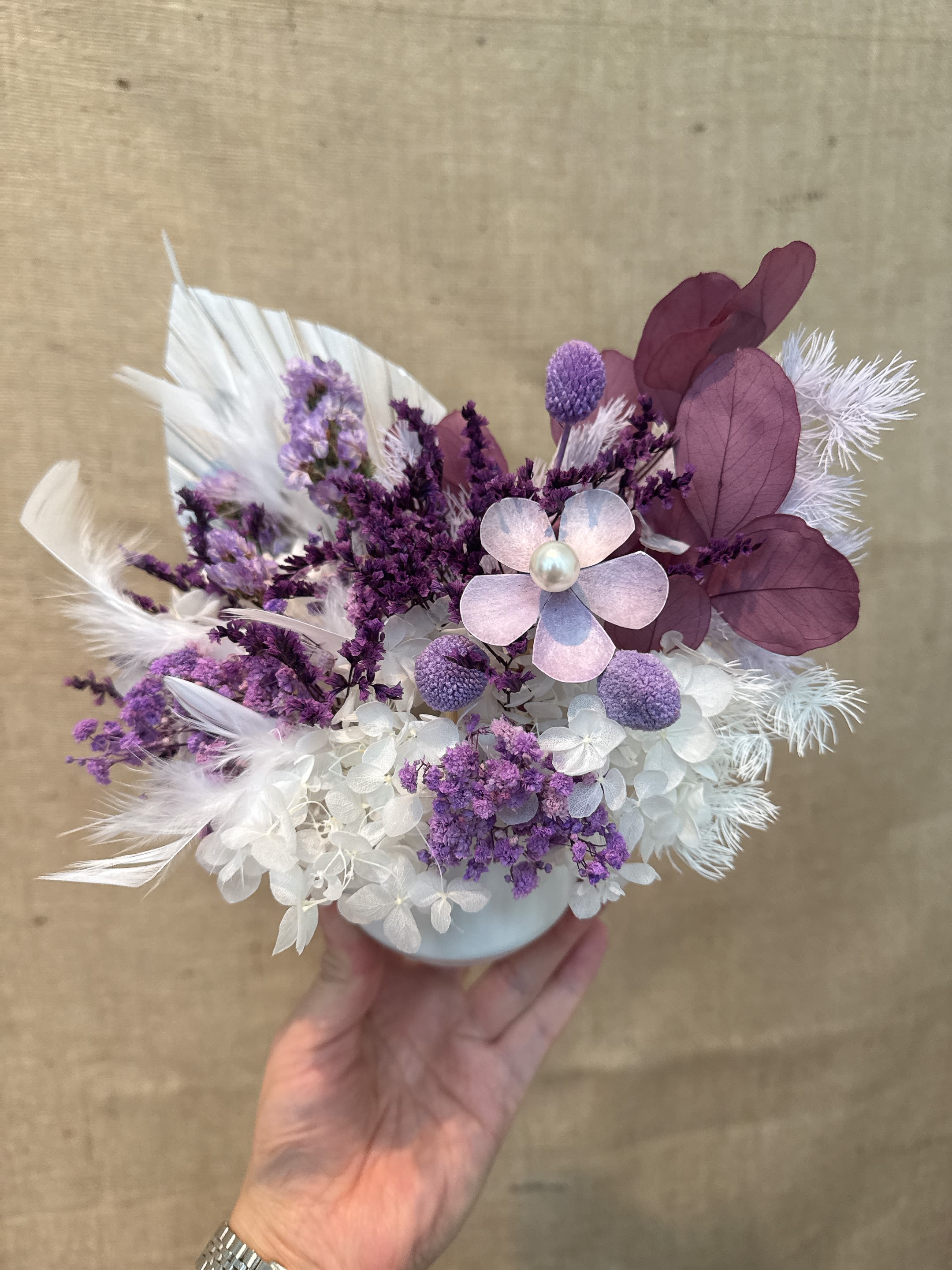 60 - This is a purple and white flower arrangement, featuring its lasting characteristics and unique texture combination. Overall style and use: This floral work has a boho or country style. Because it is an immortal flower or dried flower, it does not need maintenance (no need watering or sunlight), and can be preserved for months or even years. It is often used for home decoration, small central ornaments or as a wedding bouquet, especially suitable for occasions that want to preserve commemorative value. 