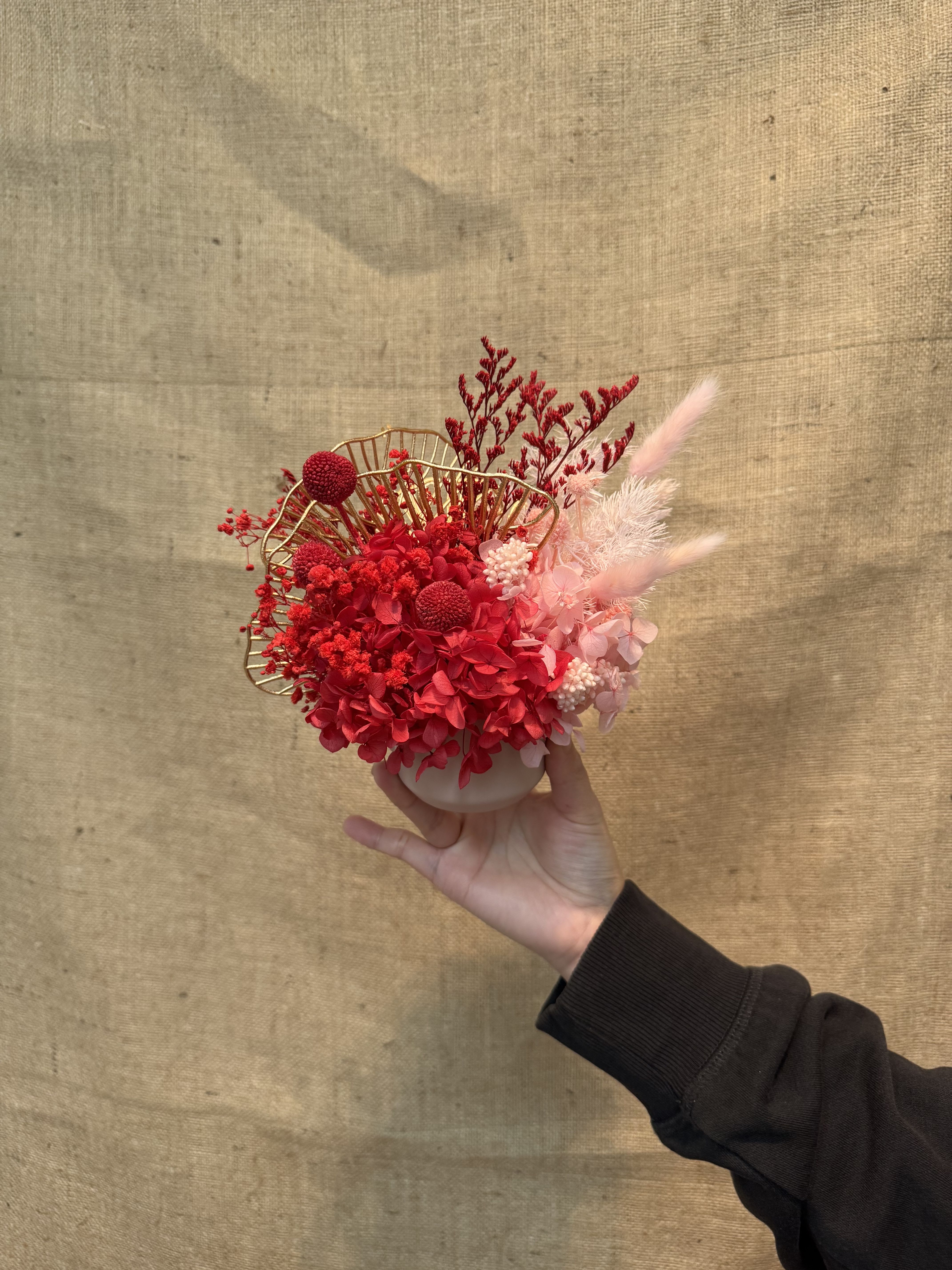 62 - This is an eternal flower or dried flower arrangement with red as the main tone, combining unique golden decoration and light pink flowers to create a strong festive atmosphere (such as the Lunar New Year). Golden decoration: The unique golden fan-shaped or mesh decoration is a golden barbed wire/wire specially designed for floral art, which is often used to enhance the festive atmosphere. The work has a strong festival and modern style, which is especially suitable for the decoration of the Lunar New Year, implying "red fire" and good luck. Because it is a dried flower or an immortal flower, it has long-lasting characteristics and is often used for home decoration, holiday ornaments or wedding decorations with specific themes. 