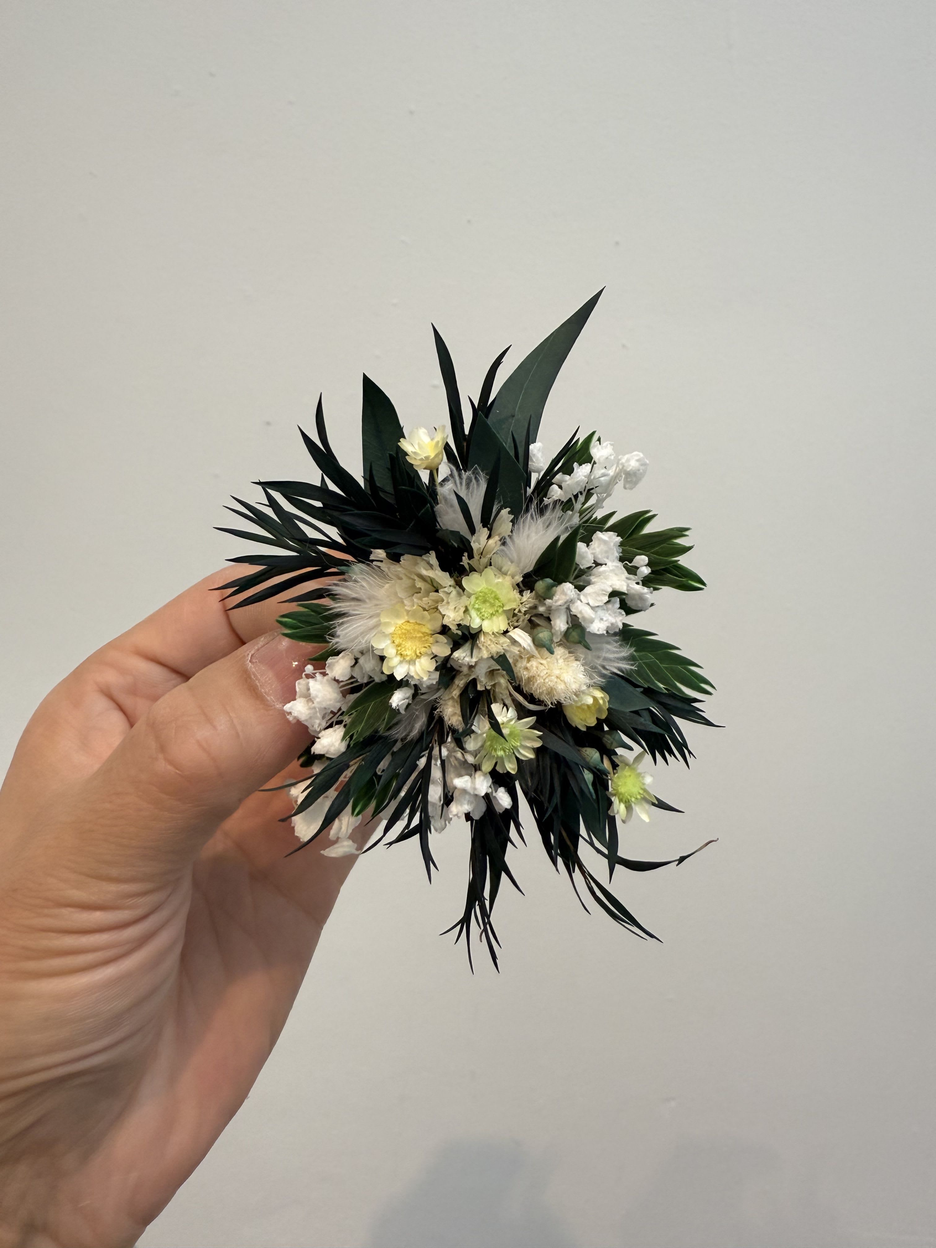 65 - This is a white, yellow and dark green permanent flower or dry flower brooch, which is often used in weddings, dances or other formal occasions. This kind of corsage is popular for its long-lasting characteristics and can be preserved for months or even years. Overall style and use: The style of this floral work is bohemian country or forest, which is very suitable for outdoor and natural weddings. It is usually used as a corsage for the groom, best man or guest, and it is pinned on the lapel of the suit. 