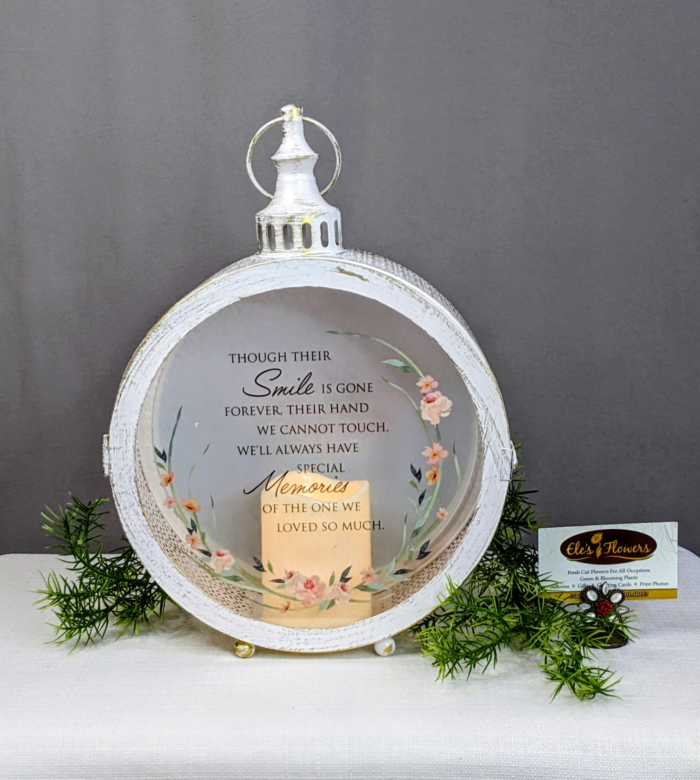 Loved So Much - Sympathy Lantern - This elegant keepsake sympathy lantern offers a gentle glow of comfort during times of loss. Framed in a soft, vintage-inspired white finish and adorned with delicate floral accents, it casts a peaceful light that honors cherished memories and enduring love. Perfect for displaying in the home or gifting as a meaningful expression of sympathy, this lantern serves as a quiet reminder that love never fades.  Printed on the glass is the touching sentiment: “Though their smile is gone forever, their hand we cannot touch, we'll always have special memories of the one we loved so much.”  A timeless keepsake meant to bring solace, reflection, and a sense of closeness when words are hard to find.   Uses 3 AAA batteries - included - for flicker candle. Indoor Use Only.  Approximate Dimensions: 15"H x 10 1/4"W x 4"D