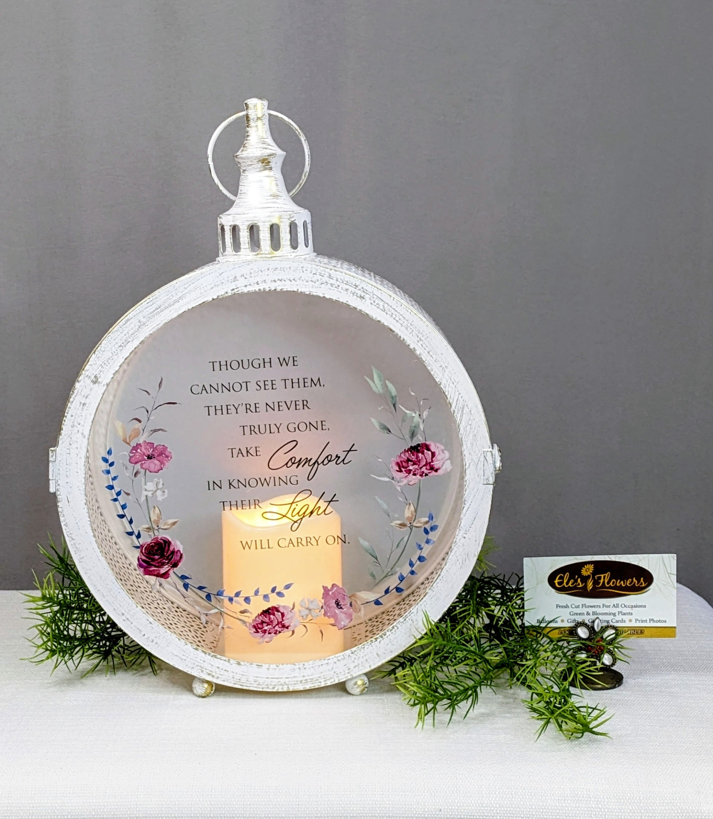 Light Will Carry On - Sympathy Lantern  - This elegant keepsake sympathy lantern offers a gentle glow of comfort during times of loss. Framed in a soft, vintage-inspired white finish and adorned with delicate floral accents, it casts a peaceful light that honors cherished memories and enduring love. Perfect for displaying in the home or gifting as a meaningful expression of sympathy, this lantern serves as a quiet reminder that love never fades.  Printed on the glass is the touching sentiment: “Though we cannot see them, they’re never truly gone. Take comfort in knowing their light will carry on.”  A timeless keepsake meant to bring solace, reflection, and a sense of closeness when words are hard to find.  Uses 3 AAA batteries - included - for flicker candle. Indoor Use Only. Approximate Dimensions: 15"H x 10 1/4"W x 4"D
