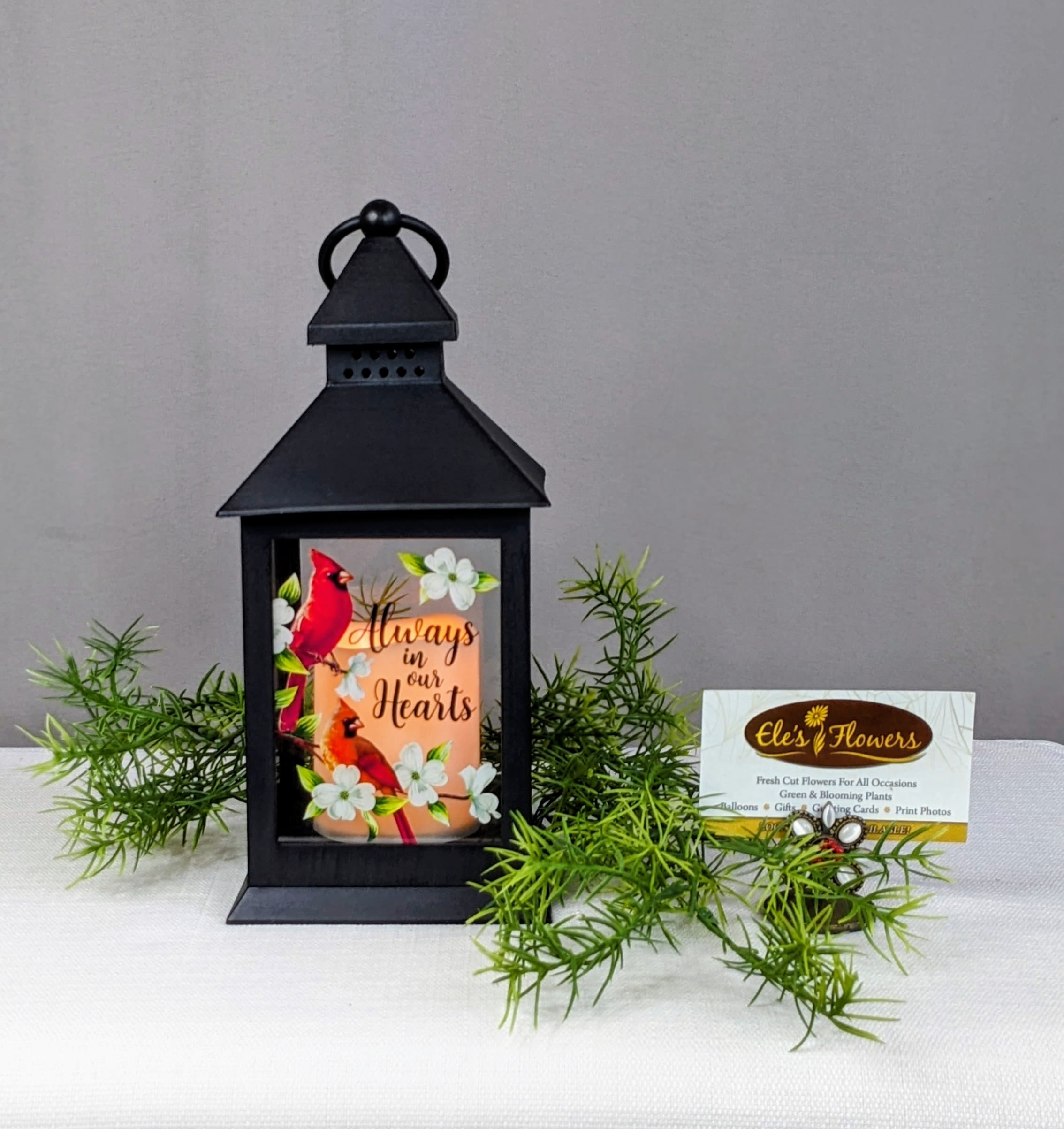 Always in our Hearts - Lantern  - A keepsake lantern with an LED flicker candle is a thoughtful memorial piece designed to provide comfort and remembrance. Made from durable plastic, the lantern features a timeless design with clear panels that allow the gentle, realistic flicker of an LED candle to shine through. This lantern serves as a lasting tribute, offering a warm and comforting glow. This lantern has the phrase, "Always in our Hearts" on the front with a cardinal and floral printed design. Indoor use only.  Approximate Dimensions: 11"H x 4"W