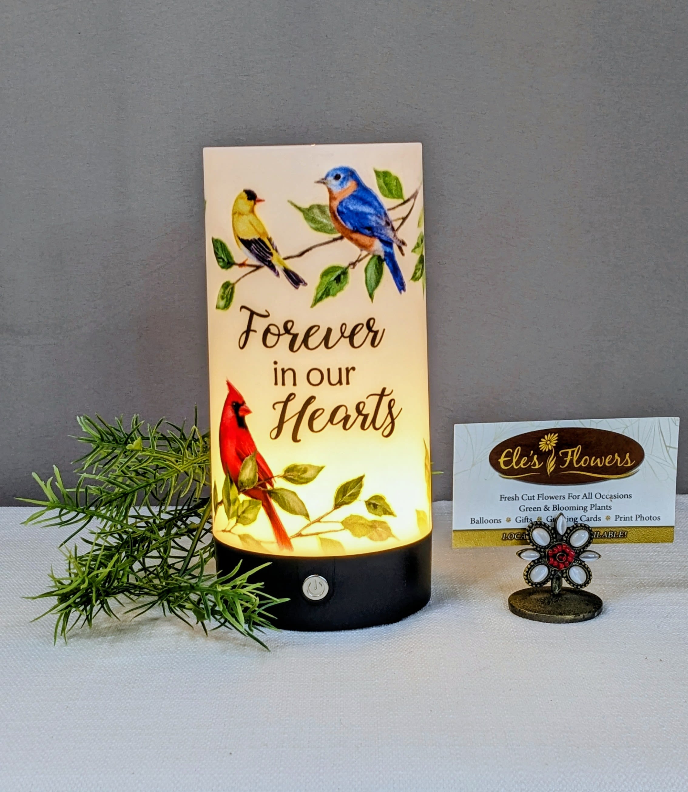 Forever in Our Hearts - Tabletop Lantern  - This lightweight tabletop cylinder lantern is a heartfelt keepsake designed to honor lasting memories with warmth and gentle light. Featuring three touch-activated lighting settings, it allows you to easily adjust the glow—from soft and subtle to bright and comforting.  Printed with the touching sentiment, “Forever in our hearts,” the lantern is beautifully adorned with a cardinal perched among leafy branches, joined by a goldfinch and a bluebird resting nearby. These beloved birds are often seen as symbols of remembrance, hope, and comfort, making this lantern a meaningful expression of love that endures. Perfect for display in the home or as a thoughtful sympathy gift, it offers a peaceful reminder that those we love are never far from our hearts.  Approximate Dimensions: 7 1/4"H x 3 1/2"D