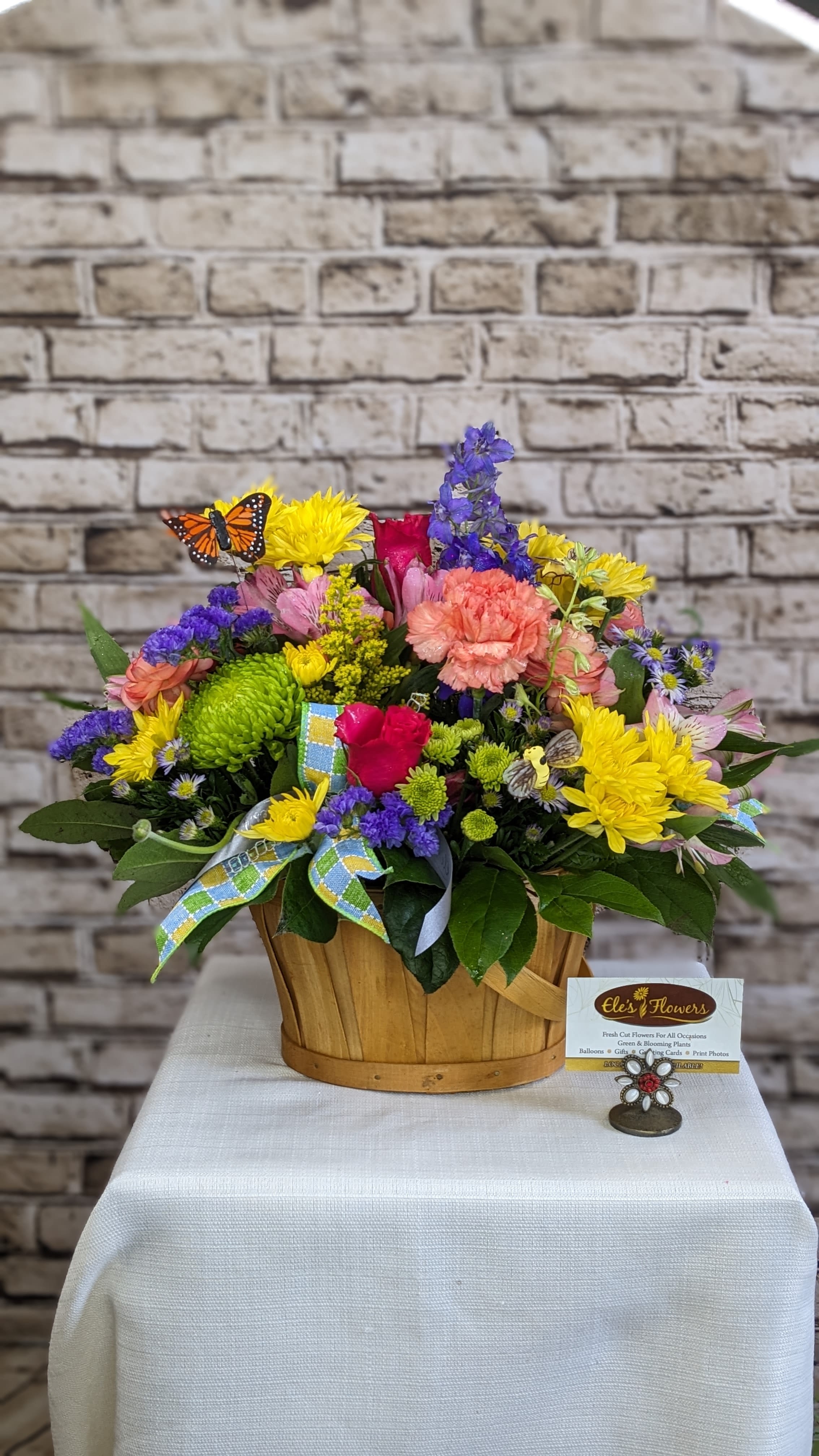 Vibrant Jubilee Basket - Celebrate the joy of the season with our Vibrant Jubilee Basket—a cheerful explosion of vibrant blooms that radiates warmth and happiness. This lively arrangement features a stunning mix of sunny yellow, pink and orange blooms, and pops of blue and purple accent flowers, all nestled in a charming wooden basket. Decorative butterflies and bees bring a whimsical, garden-fresh feel, while a plaid pastel bow adds the perfect finishing touch. Perfect for any occasion, this arrangement is a burst of seasonal delight.   Blooms may vary from photo.    Approximate Dimensions: 17"H x 14"W