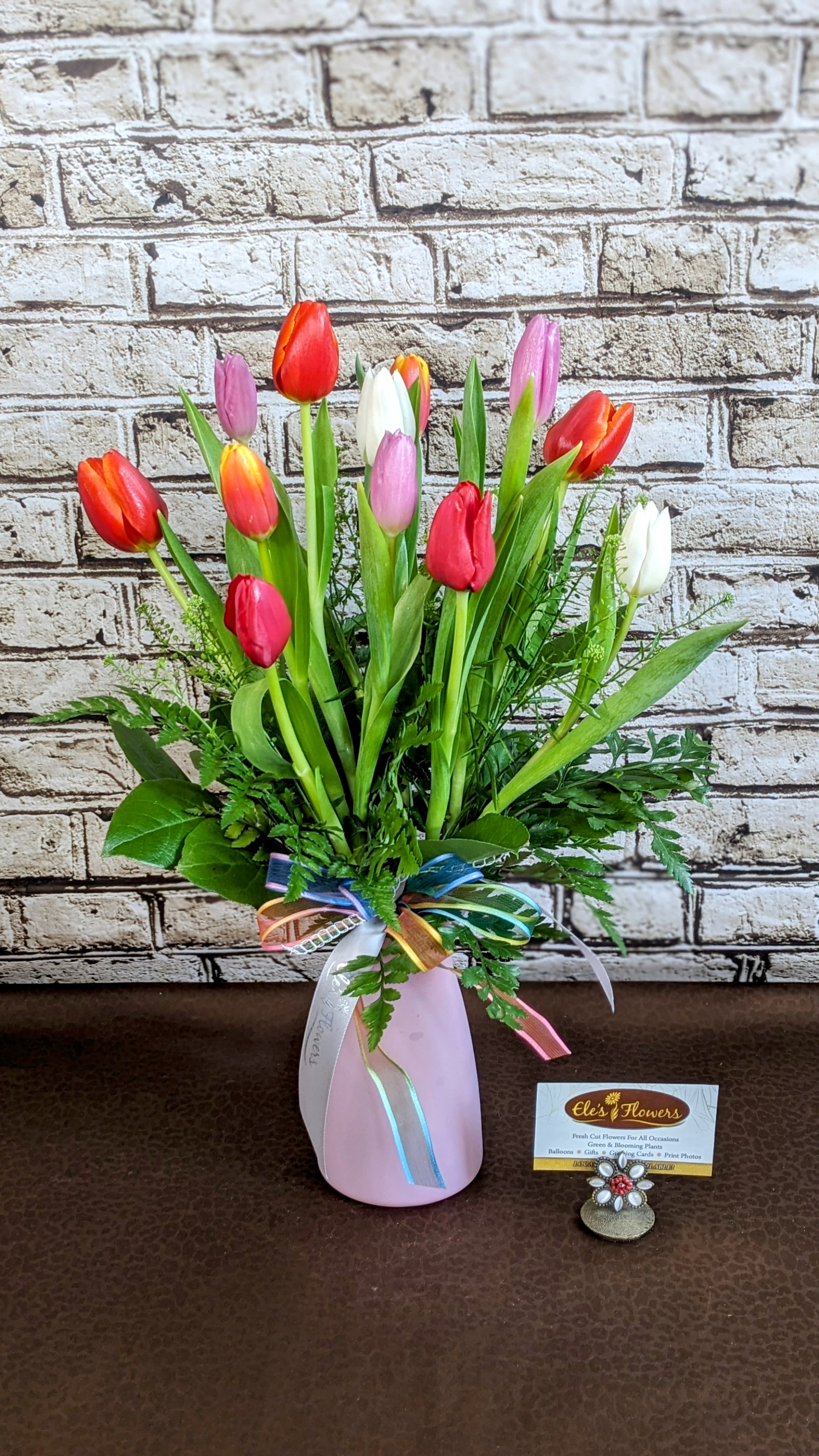 Tulip Bliss - An arrangement of a dozen mixed-color tulips, artfully paired with fresh greenery and delicate accent flowers. This cheerful and elegant bouquet is a perfect way to brighten someone’s day with a touch of charm and spring cheer! Colors may vary from photo  We source the freshest flowers available to craft a stunning floral arrangement tailored to the recipient's preferences. If you have any specific requests, please include them in the 'special instructions' section of your order to ensure we bring your vision to life. Otherwise, our talented designers, Brett and Sara, will expertly handle the details for you.  Standard Size is an excellent desktop size or small table. Approximate Dimensions: 18”H x 16”W 