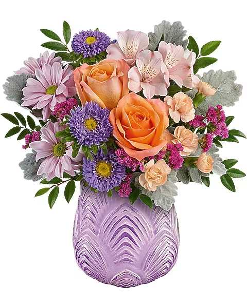 Teleflora's Brilliant Purple Bouquet - This fun arrangement bursts with springtime energy, beautifully showcased in a sculpted glass vase with a soft lavender hue and frosted finish, evoking the essence of a blossoming garden. This bouquet showcases peach roses, light pink alstroemeria, miniature peach carnations, lavender Matsumoto asters, and lavender daisy spray chrysanthemums, all beautifully complemented by raspberry statice, huckleberry, and dusty miller. This bouquet is elegantly arranged in the Dreamy Blooms vase.   Orientation : All-Around  All prices in USD ($)  Due to an increase in the cost of roses, some product prices may be impacted during the Valentine’s delivery period.