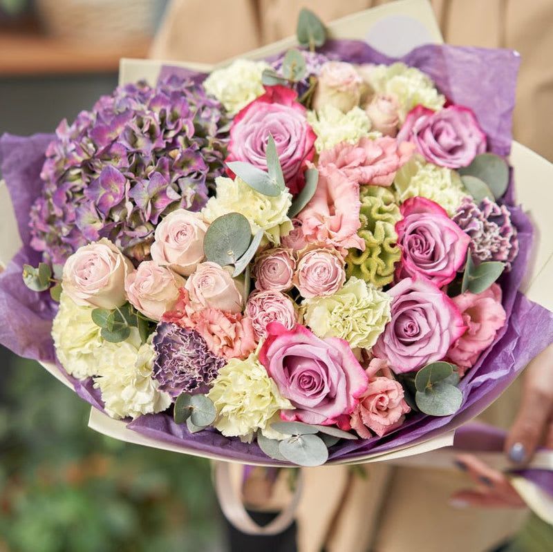 Delicate To the Core - This arrangement is a harmonious blend of soft pastels and rich, earthy tones, exuding elegance and natural charm. At its core, lush purple hydrangeas command attention with their bold, rounded blooms, setting a regal tone. Surrounding them, light pink lisianthus adds an airy and delicate touch, while soft pink roses and spray roses bring classic romance and a refined sweetness.  The unexpected addition of green and purple carnations introduces a playful yet sophisticated texture, creating a dynamic interplay of colors that feels both fresh and timeless. Accentuated by the silvery-green foliage of eucalyptus, the arrangement gains an organic flow, adding depth and a hint of fragrance to the design.  Perfect for occasions that call for understated beauty and refinement—whether as a centerpiece, a gift, or a heartfelt gesture—this bouquet offers a delightful mix of textures and hues. Its seamless balance of soft florals and lush greenery creates a versatile arrangement that conveys grace and thoughtful sentiment.