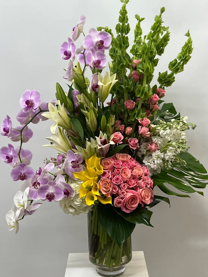 Opulence Masterpiece  - Envision opulence at its finest with our super luxury flower arrangement, a true masterpiece for the discerning connoisseur. This extravagant display combines the grace of orchids, the purity of lilies, the vibrancy of bright yellow mini calas, and the romance of pink roses, all harmoniously adorned with lush monsteras leaves. Housed in a tall crystal vase, this stunning ensemble exudes sophistication and grandeur, making it a captivating centerpiece that will elevate any space with its unparalleled beauty and charm.