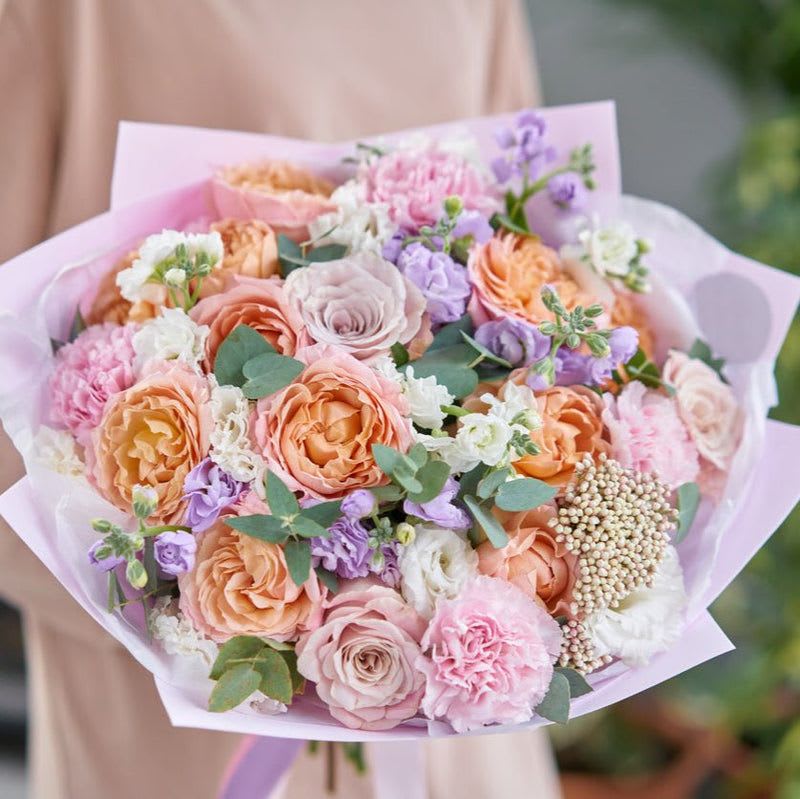 Pastel Pop - Introducing a breathtaking hand-tied bouquet that blends elegance and charm in a delightful array of colors and textures. This stunning arrangement features beautifully light lavender stock, soft peach roses, enchanting lavender roses, and delicate light pink carnations, accentuated by the ethereal presence of white lisianthus and the refreshing touch of baby blue eucalyptus.  The light lavender stock provides a lovely base, its subtle fragrance enhancing the bouquet's allure. Soft peach roses add warmth and a romantic touch, symbolizing gratitude and admiration. The lavender roses offer a unique twist, combining beauty with a hint of mystique, while the light pink carnations bring an element of joy and playfulness. The white lisianthus brings in a layer of elegance, creating a harmonious blend of shapes and colors.  Topping it all off, the fragrant baby blue eucalyptus creates a lovely contrast, adding a soothing pop of color and a refreshing aroma. Hand-tied with care, this bouquet is lovingly wrapped in natural materials, making it a perfect gift for birthdays, anniversaries, or any special occasion. With its captivating colors and exquisite floral composition, this hand-tied bouquet is sure to bring smiles and joy to anyone who receives it.