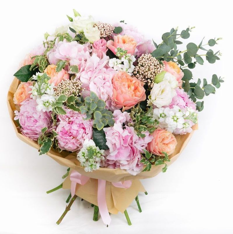 Peony Impressions - This exquisite hand-tied bouquet embodies a romantic and elegant charm, perfect for any special occasion. At its heart, lush garden roses in soft pinks and vibrant coral hues provide a captivating focal point, emitting a delicate fragrance that enhances its allure. Surrounding these roses, fluffy soft pink hydrangeas add a touch of whimsy and fullness, creating an inviting texture.  Delicate stock flowers in complementary shades bring a subtle sweetness, while the elegant lisianthus contributes a refined and gentle touch with its graceful bell-like blooms. The addition of airy rice flowers introduces a lightness to the arrangement, enhancing its romantic feel.  A curated selection of elegant greens serves as the perfect backdrop, with assorted foliage that adds depth and contrast. Varieties of lush ferns, rich eucalyptus, and dainty sprigs intertwine, giving the bouquet a sophisticated finish.  This hand-tied arrangement is not just a bouquet; it's a stunning expression of beauty and elegance, ideal for weddings, anniversaries, or as a heartfelt gift to someone special. Each bloom is thoughtfully arranged to showcase its unique beauty, creating a harmonious blend of colors and textures that will leave a lasting impression.