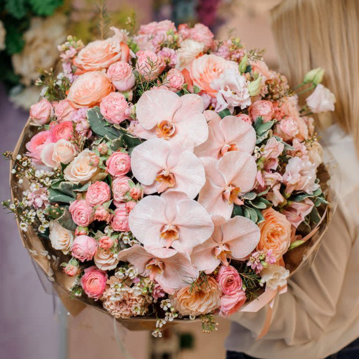 Princess Peach - This exquisite bouquet is a true celebration of soft, harmonious hues and elegant textures, showcasing a delicate blend of peachy-pink spray roses and full-bodied regular peachy-pink roses. The arrangement dances with gentle layers of romantic roses, each bloom adding its own unique charm. At the heart of this beauty, phalaenopsis orchids sit gracefully, their smooth, satin-like petals exuding a refined sense of luxury and tranquility. Their creamy white tones are accented with a touch of lavender, creating a striking contrast against the warm, coral pinks of the roses.  A scattering of wax flowers adds a whisper of airy freshness, its tiny, star-shaped blooms contributing a subtle, ethereal quality to the bouquet. For an extra layer of delicacy, lisianthus flowers gently peek through, their soft ruffled edges echoing the romance of the roses and orchids, and infusing the bouquet with a soft, almost ethereal beauty.  This luxurious arrangement is a perfect choice for those who appreciate a balanced, yet bold, mix of texture and color. Its dreamy composition is ideal for weddings, anniversaries, or any occasion where a touch of refined elegance is desired. Each flower within this bouquet is carefully selected to create a sophisticated yet natural feeling, making it an unforgettable floral masterpiece.