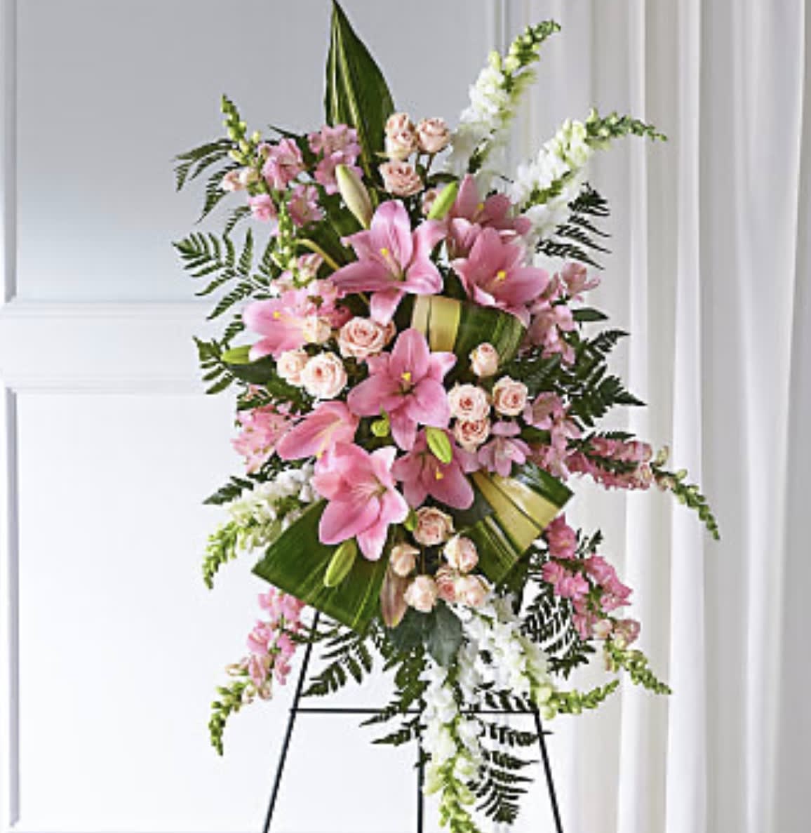 Elegant Embrace Standing Spray - ELEGANT EMBRACE STANDING SPRAY  Our Elegant Embrace Standing Spray shares the warmth and comfort your loved ones need during this difficult time through each blushing bloom. A local florist beautifully combines lilies, spray roses and snapdragons with greens to express your heartfelt love.   SUBSTITUTION POLICY Always deliver the freshest flowers! Please note the bouquet pictured reflects our original design.  If the exact flowers or container in this arrangement are not available, our local florists will create a beautiful bouquet with the freshest available flowers.