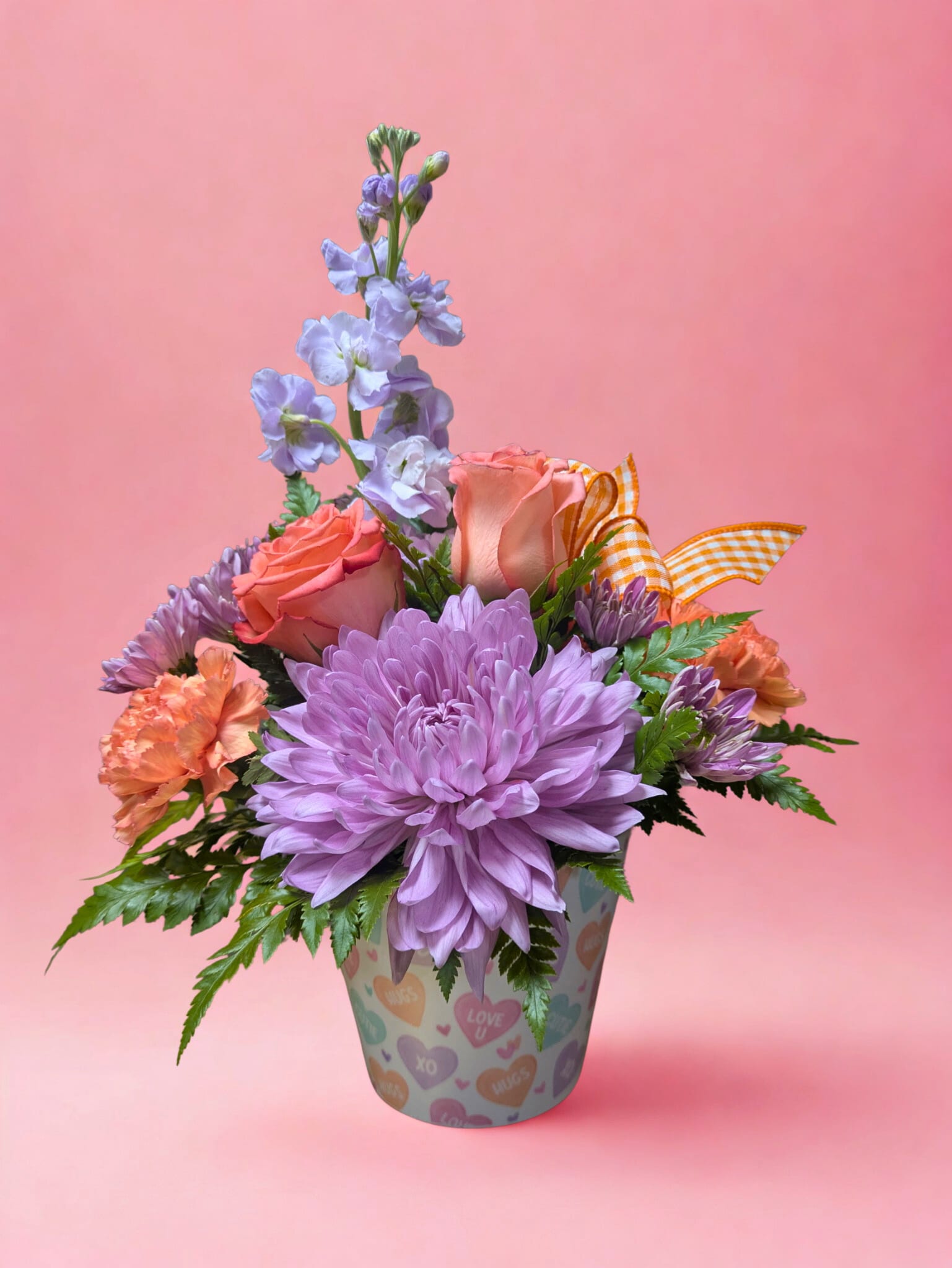 Sweet Talk - Playful, colorful, and full of Valentine’s Day charm, Sweet Talk is a bouquet that says it all without words. Arranged in a whimsical container adorned with candy hearts, this lively mix features soft lavender cremone, a warm coral rose, cheerful orange carnations, plush lavender cushions, and fragrant lavender stock, finished with a bright orange plaid ribbon for a fun pop of color.  Perfect for sending a flirty “thinking of you” or a heartfelt “I love you,” this arrangement is a sweet, vibrant way to make someone’s heart smile.   All flowers, floral colors, and containers are subject to change due to availability.