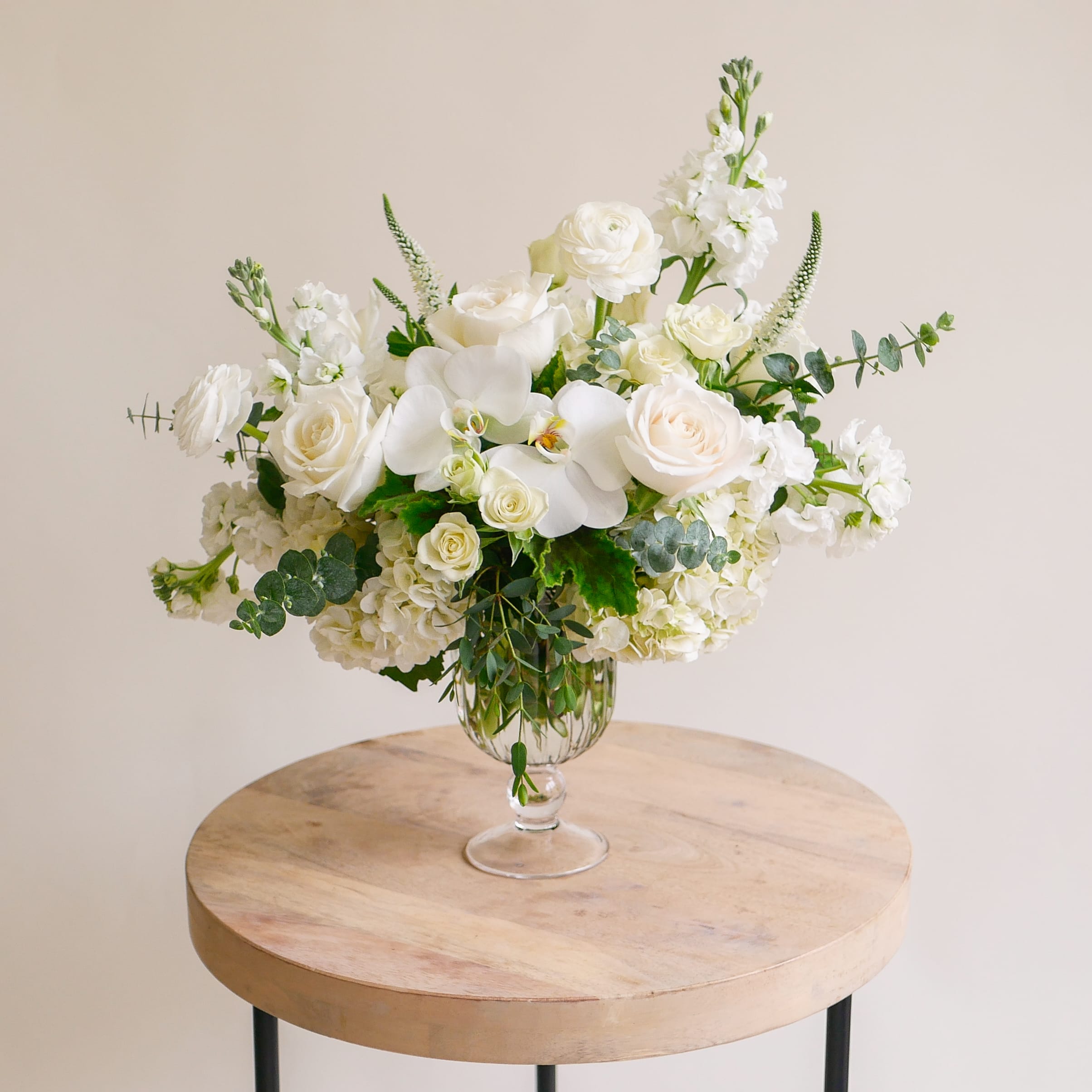 Beverly Butterfly - This ethereal arrangement showcases an organic selection of classic white blooms including roses, ranunculus, stock, orchids, hydrangeas and more. Designed within a premium goblet vessel, this arrangement approximately measures 16" tall and 16" wide.