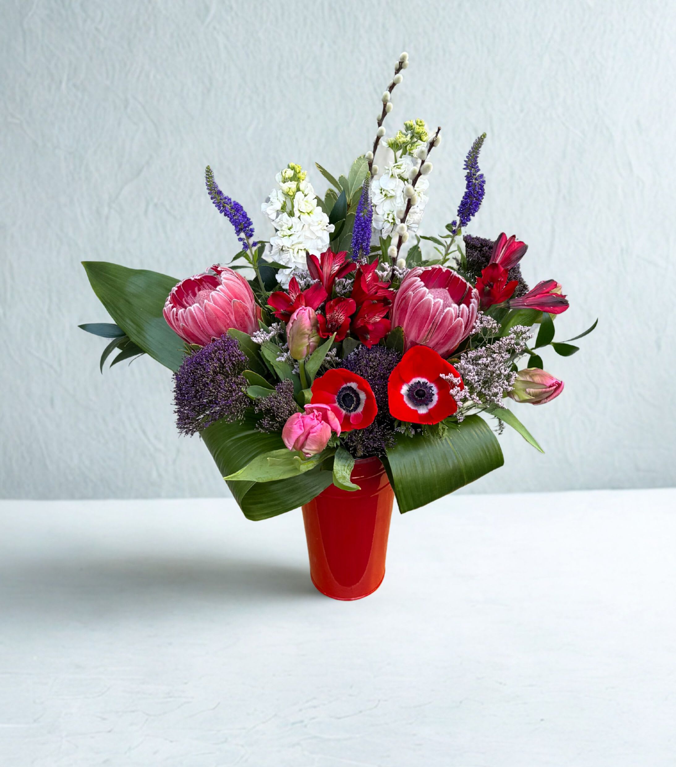 Hillsides Scarlet Devotion - A bold Valentine’s arrangement featuring rich red blooms, fresh greenery, and elegant texture, designed in a striking red vase to beautifully express love and passion.