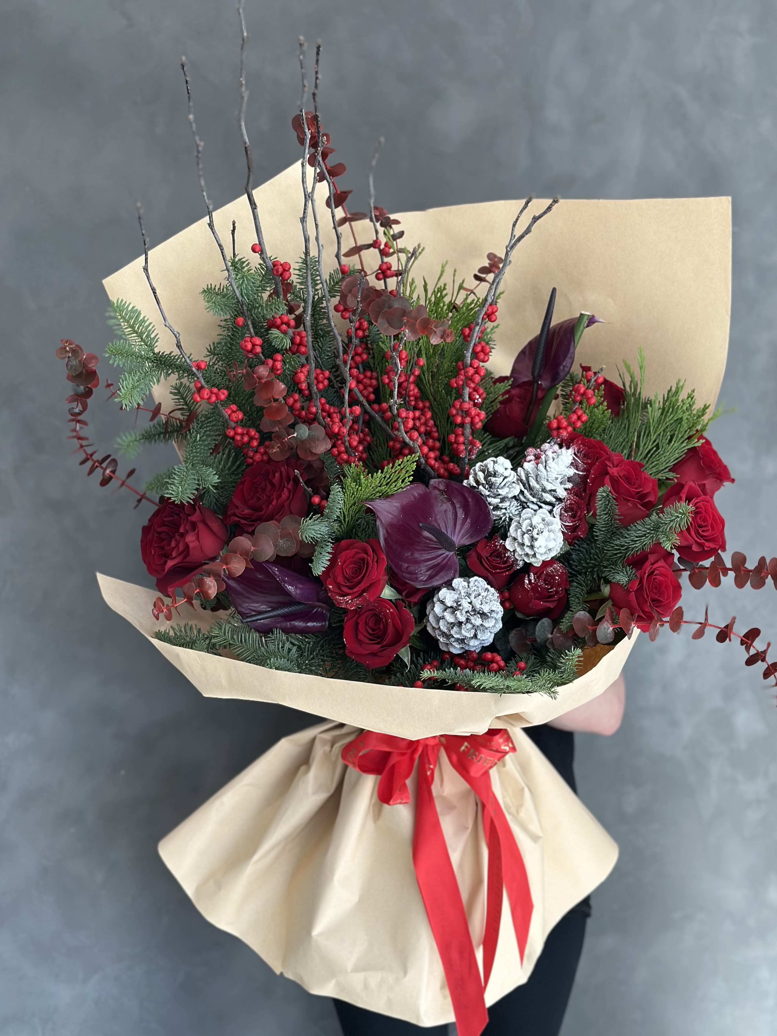 Frosted Wishes - This lush, winter-inspired bouquet combines the vibrant red of roses with the rich textures of pine cones, winter berries, and spruce branches, accented by the striking elegance of anthurium. Perfect  for the season, it brings together a blend of festive warmth an natural beauty, evoking cozy, holiday charm.