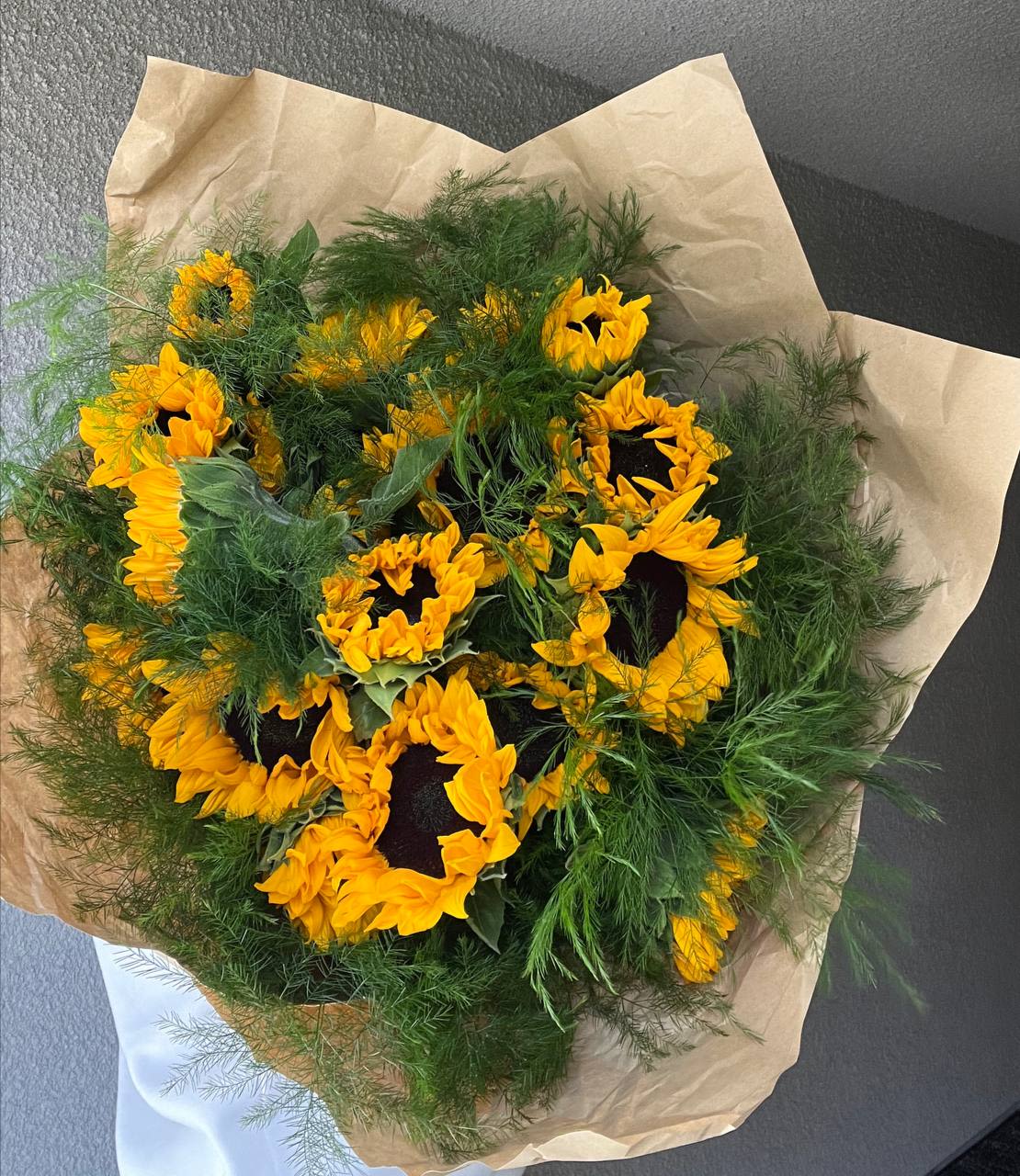 Sunlight - Bouquet “Sunlight” is a true embodiment of warmth, joy, and the energy of nature.  Bright sunflowers with golden petals shine like little suns, radiating light and positivity. Their deep black centers create a striking contrast, adding depth and expressiveness to the composition. The soft, feathery greenery brings freshness and lightness, evoking the feeling of a summer breeze dancing across a blooming field. The natural kraft paper wrapping enhances the bouquet’s organic aesthetic, making it a warm and cozy accent.  “Sunlight” is the perfect gift for those who deserve a touch of happiness, inspiration, and sunshine in their day.