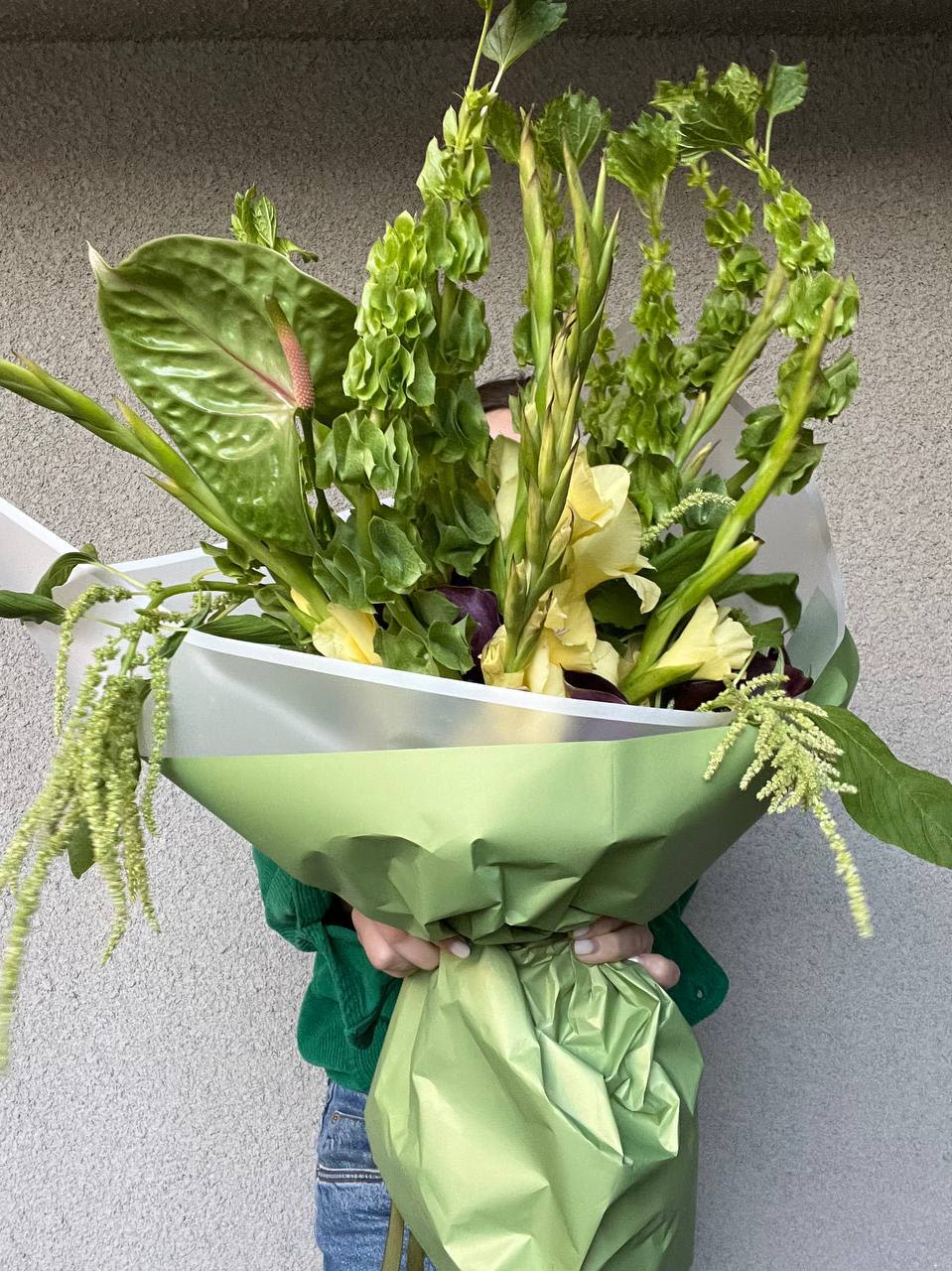 Emerald Forest - Bouquet “Emerald Forest” is a true embodiment of natural harmony and freshness, inspired by wild woodland landscapes.  Its composition is dominated by rich green hues: airy bells of molucella resemble tiny forest chimes, while striking anthuriums with glossy leaves add an exotic touch. Soft cream-yellow gladioli bring lightness and delicacy, balancing the deep greenery. Graceful cascading elements, such as amaranthus, create a sense of fluidity and natural movement, reminiscent of trailing vines in a tropical forest.  This bouquet is a perfect choice for those who appreciate natural aesthetics, unconventional combinations, and the enchanting mystery of a lush, untamed forest.