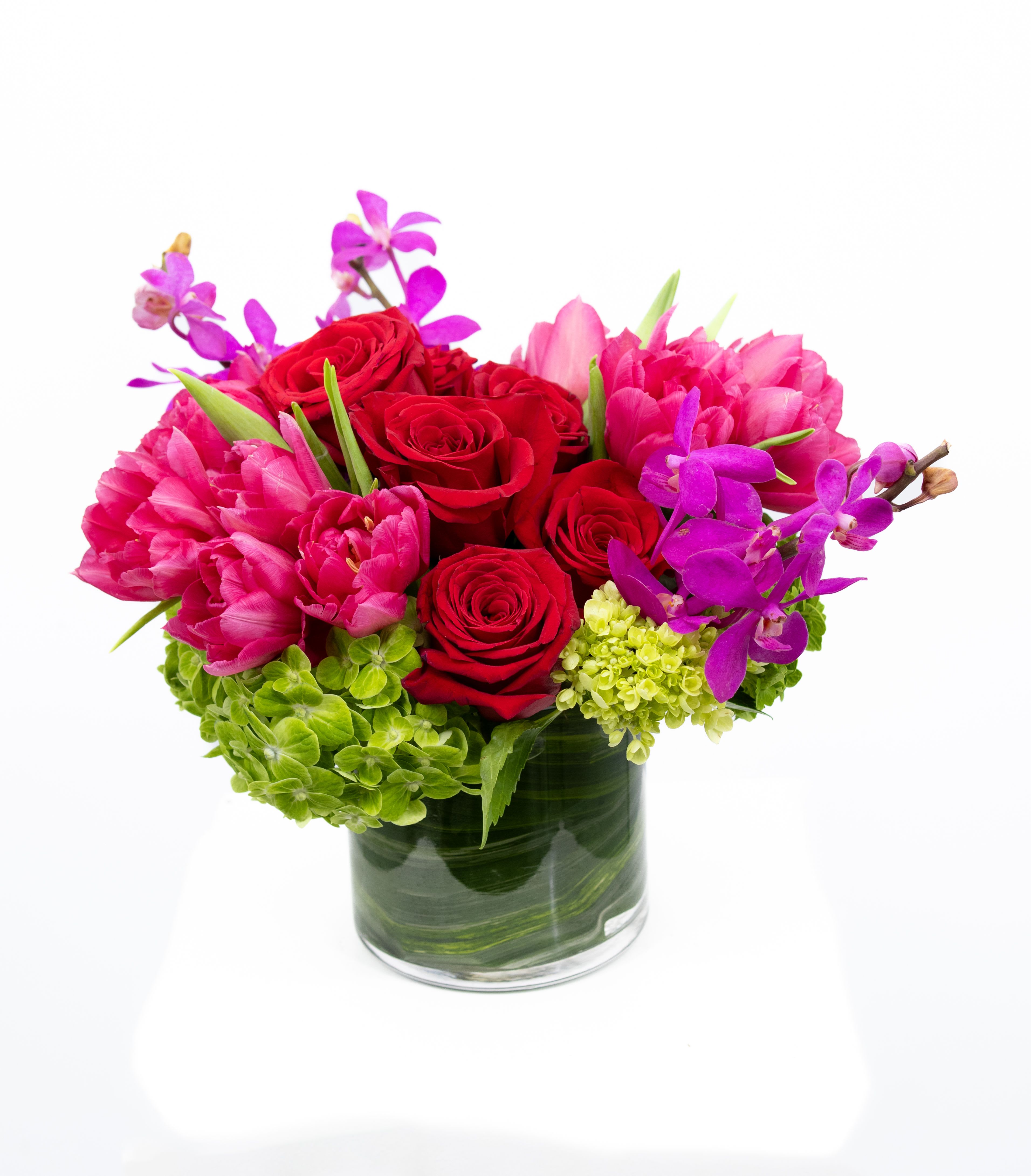 Hope - A vibrant floral arrangement with red roses at the center, surrounded by bright pink tulips, purple orchids, and green hydrangeas. The flowers are arranged in a clear glass vase lined with lush green leaves.