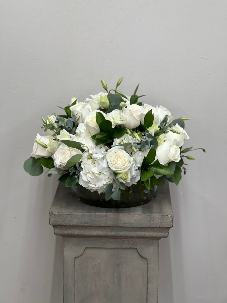 Bliss - Simple and elegant white hydrangeas, roses and lisianthus accented with different greenery and arranged in a large, low cylinder vase.   Flower shades may vary depending on variety. 