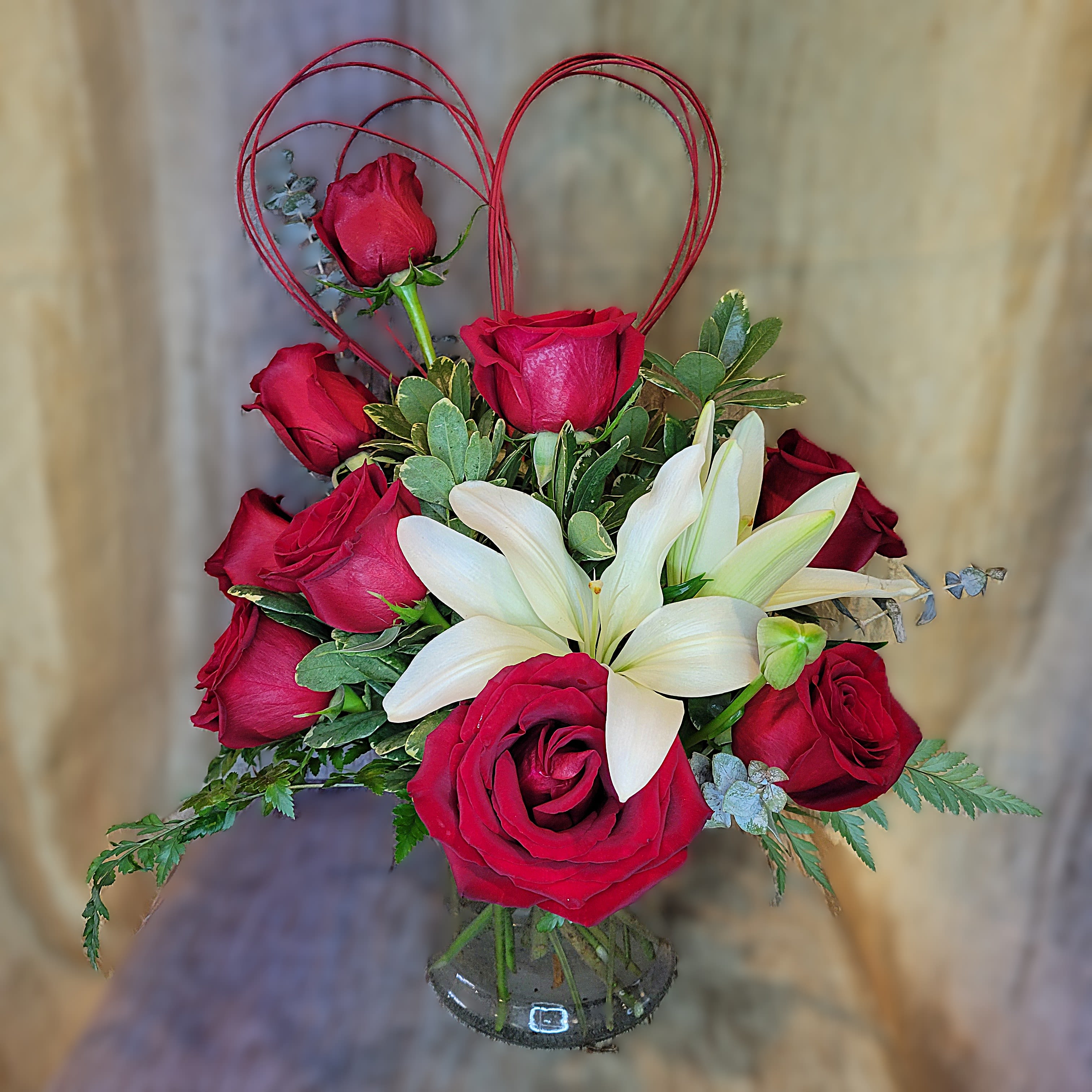 Refined Romance - PREORDER - The classic combination of Red Roses and Lilies accented with charming heart twist!  Give a touch of playful elegance to your refined Valentine.  Lily colors may vary.  PICTURED AT DELUXE SIZE  Available after 02/06/2026