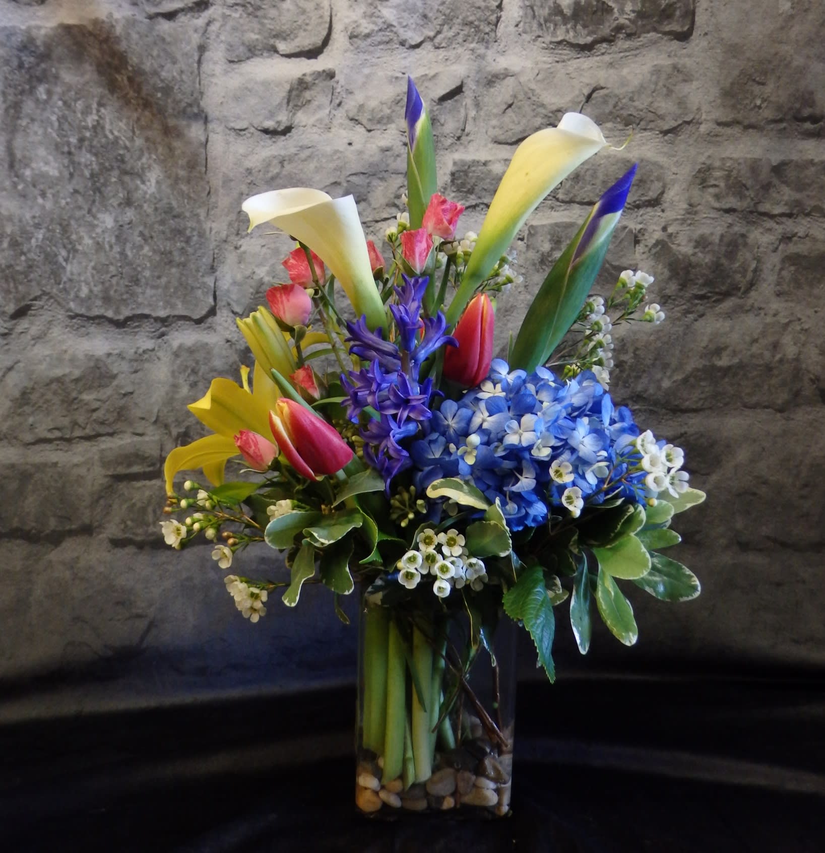 Spring Squared - "Spring Squared" is a lively burst of seasonal color, artfully arranged in a modern square vase. Featuring bold blue hydrangea, sunny yellow lilies, elegant calla lilies, tulips, and delicate accents, this design captures the fresh, uplifting spirit of spring in a stylish and cheerful display. 