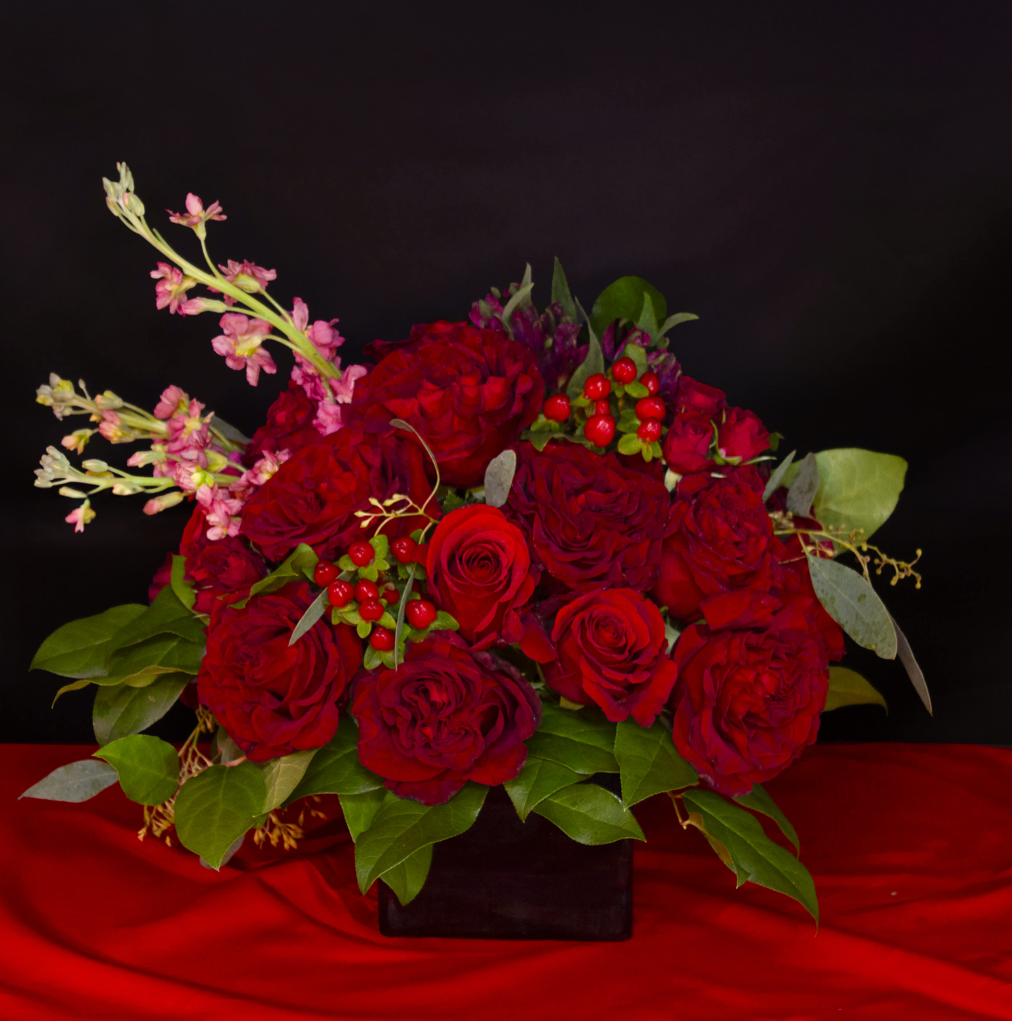 'Three Shades of Red' by Madison's Flowers - Fiery, passionate reds explode from lush glossy greens, and accents of seeded eucalyptus, in a sleek black glass cube. Modest Mauve stock springs lovingly from a bed of deep Sexy Red, Heart Roses, and red hypericum berries. In colors so vivid your cheeks will blush every shade of red. Send some passion today. A Madison Flowers original design. Vase is 5x5x5