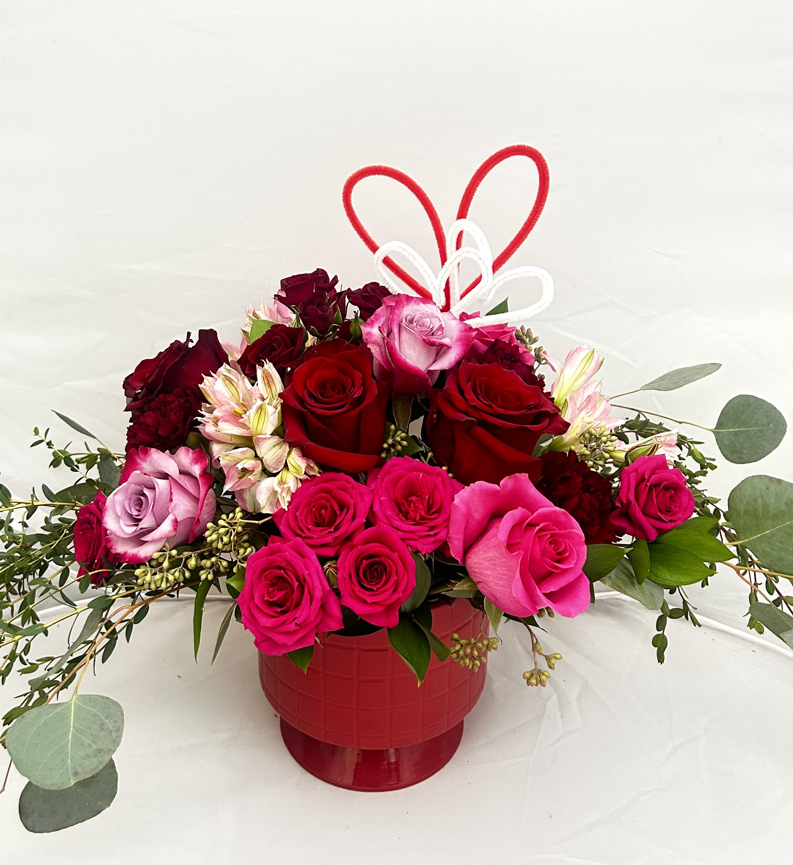 Wild For You - Express your affection with the Wild For You arrangement. This romantic display features a lush mix of deep red and hot pink roses, accented with delicate alstroemeria and sprigs of eucalyptus. Set in a vibrant red keepsake vase and topped with a whimsical heart-shaped wire detail, it’s a perfect gift for anniversaries, Valentine's Day, or any moment you want to share some love.