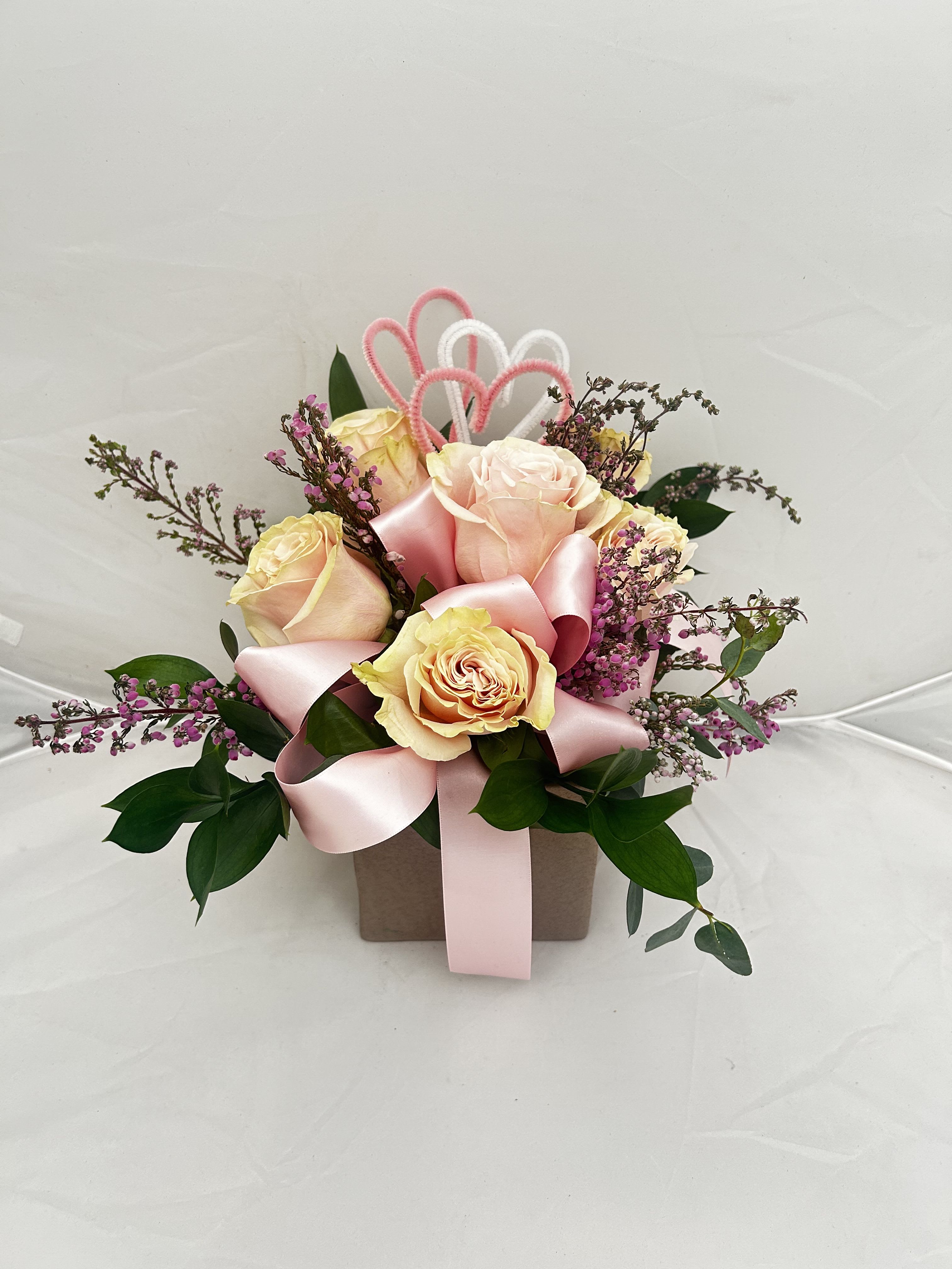 Valentine Garden Of Roses - Enchant that special someone with the Valentine Garden of Roses. This sweet and compact arrangement features soft, bi-color peach and pink roses paired with delicate purple waxflower and lush greenery. Topped with a charming pink ribbon and twin heart accents, it’s a lovely way to share your heart.