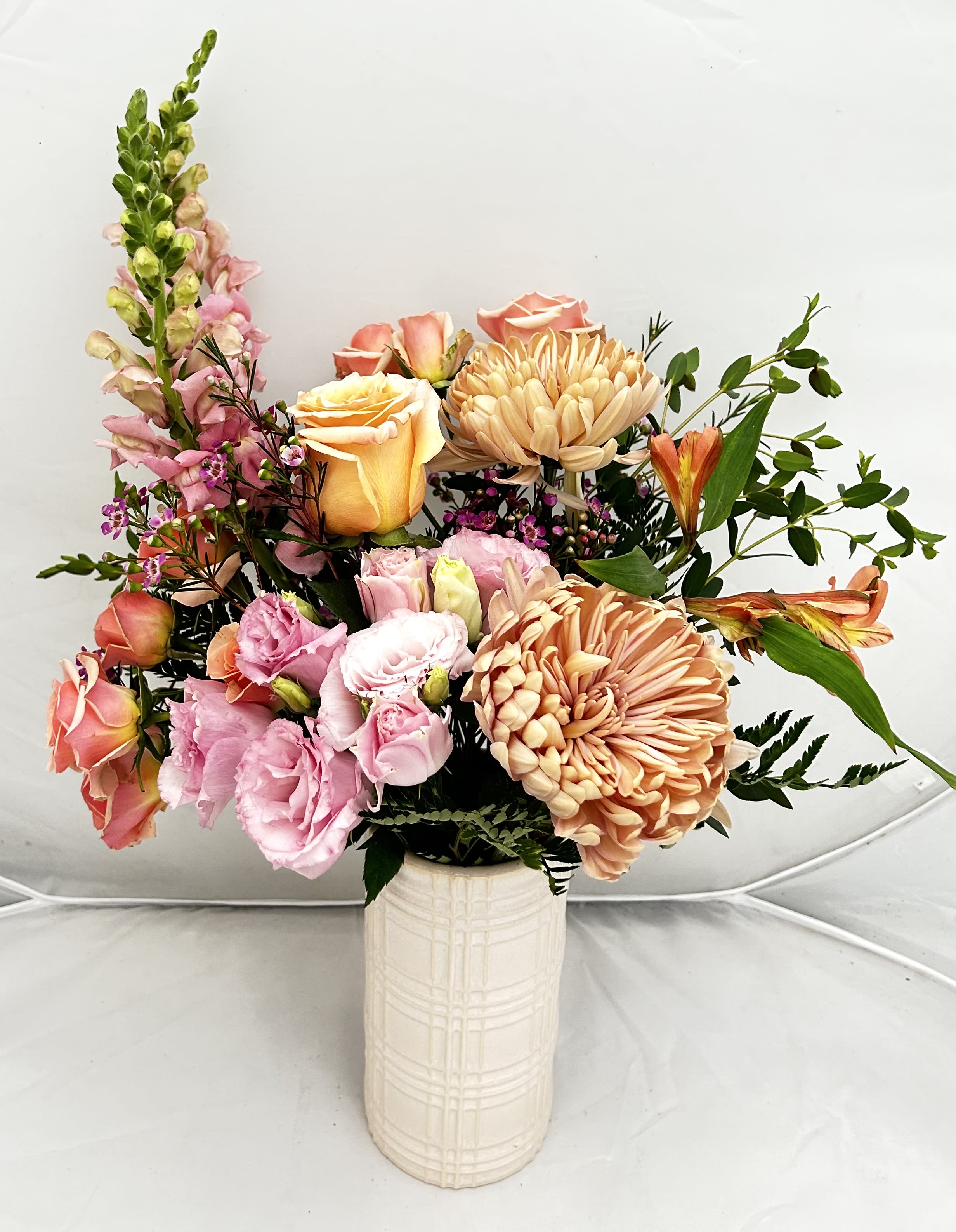 Just Peachy - Brighten any space with our Just Peachy arrangement, a lush and garden-inspired display. This design features a stunning blend of peach roses, vibrant pink spray roses, and elegant snapdragons, complemented by textured chrysanthemums and airy greenery. Presented in a modern, cream-colored ceramic vase with a subtle grid pattern, it’s a sophisticated choice for birthdays, "just because" moments, or seasonal home decor.