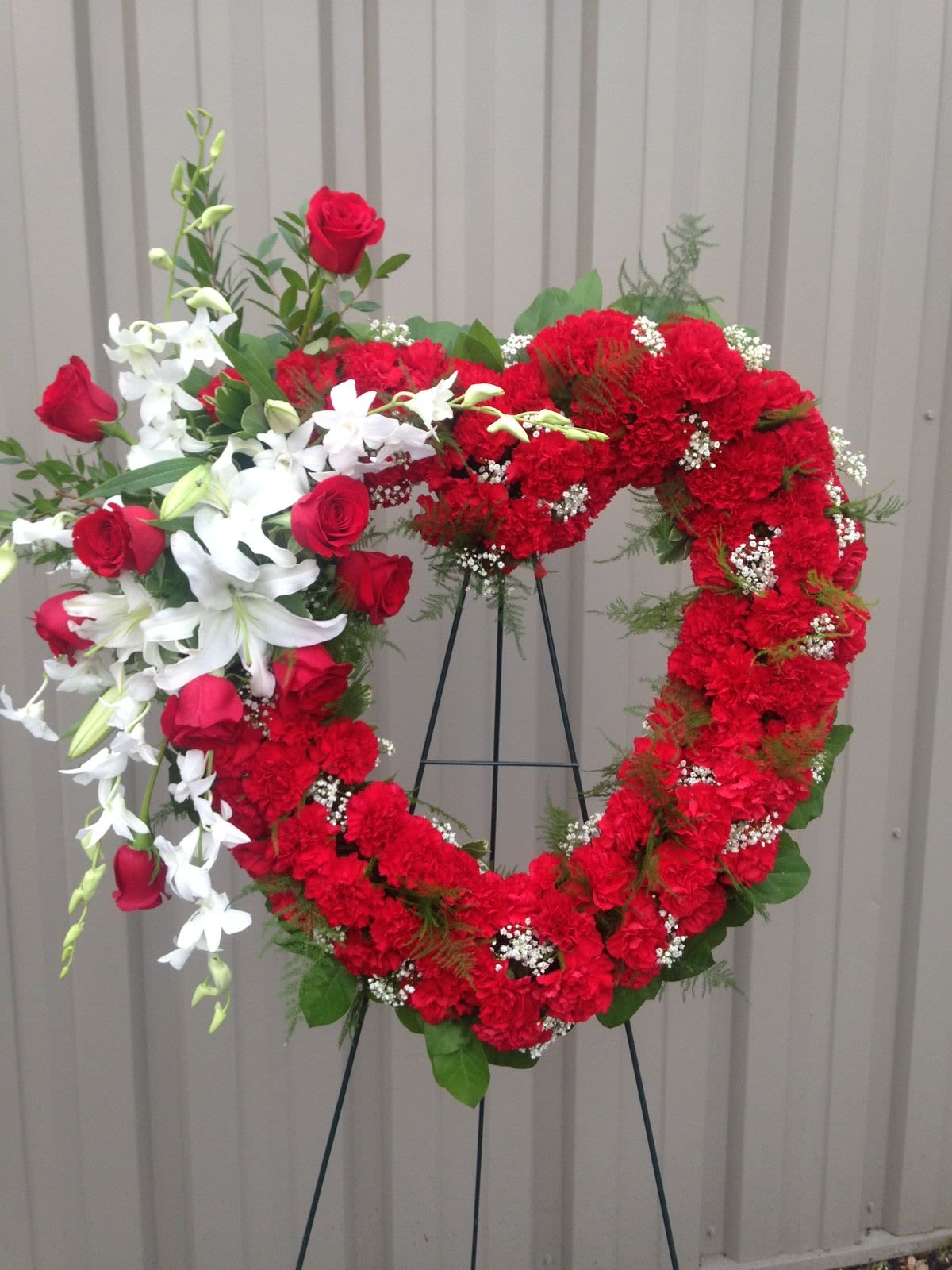 Red Heart - This standing arrangement is created to lend comfort during a difficult time. This sympathy arrangement is created with red carnations, red roses, white lilies and dendrobium orchids stems.
