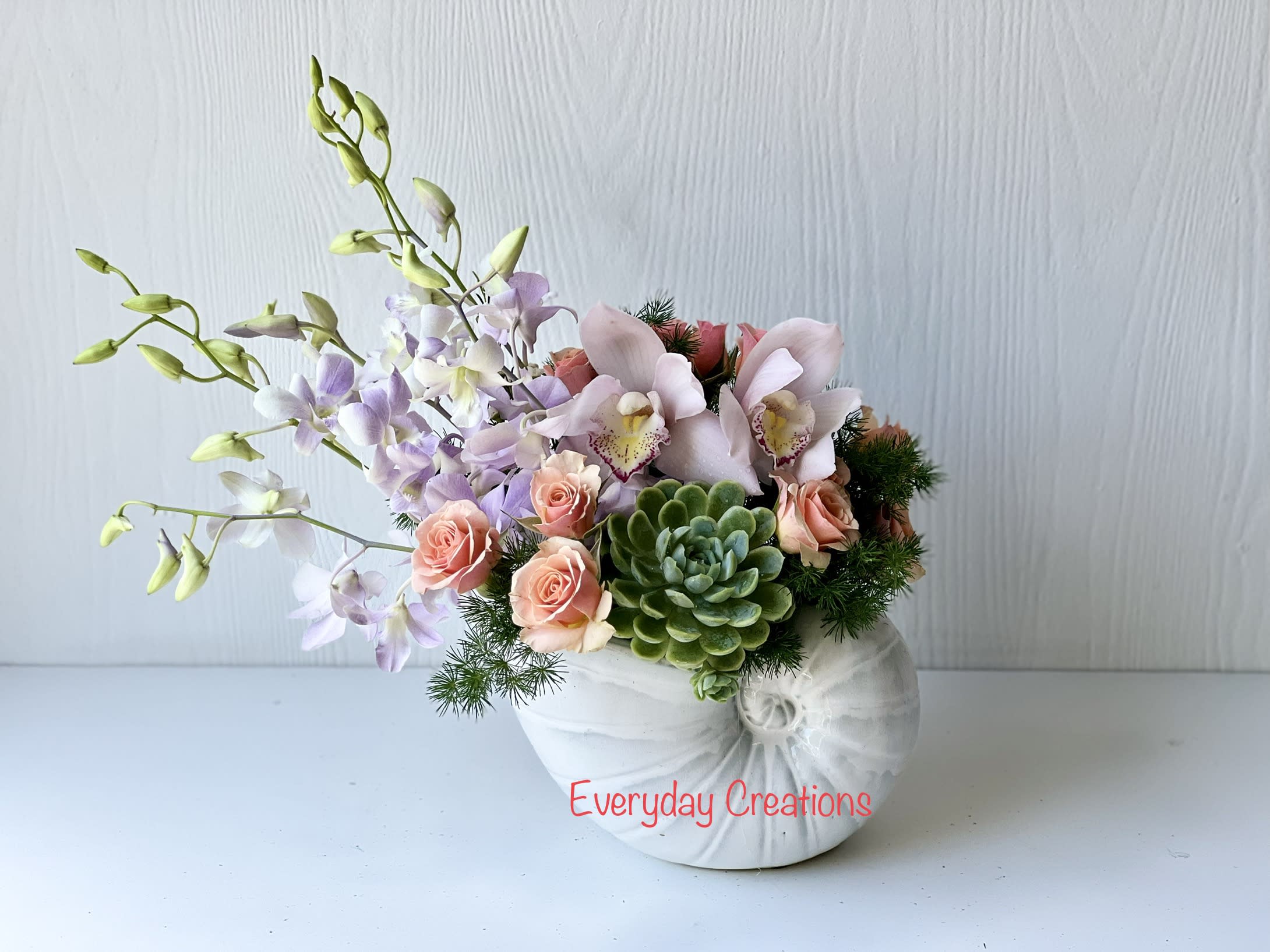 EC86 -  Coastal Charm  - Bring the tranquility and beauty of the beach into your space with this arrangement. This luxurious beachy floral arrangement features a stunning selection of premium flowers, including orchids, tulips, spray roses, succulents and a variety of greenery. All of the blooms are expertly arranged in a beautiful and unique shell vase to bring a sense of coastal charm.  Arrangement size: approx 10" wide, 13" high
