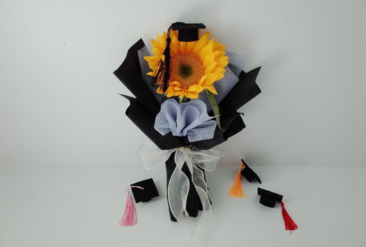 Graduation Glow — Sunflower Edition - Celebrate their big moment with a burst of sunshine.   This cheerful sunflower bouquet is wrapped in soft pastel tones and topped with a tiny graduation cap — a sweet symbol of achievement, growth, and new beginnings. Whether it’s for a friend, sibling, or student, this bouquet delivers warmth, pride, and joy in one perfect bloom.  A small gift with a big message: You did it, and the future is bright.