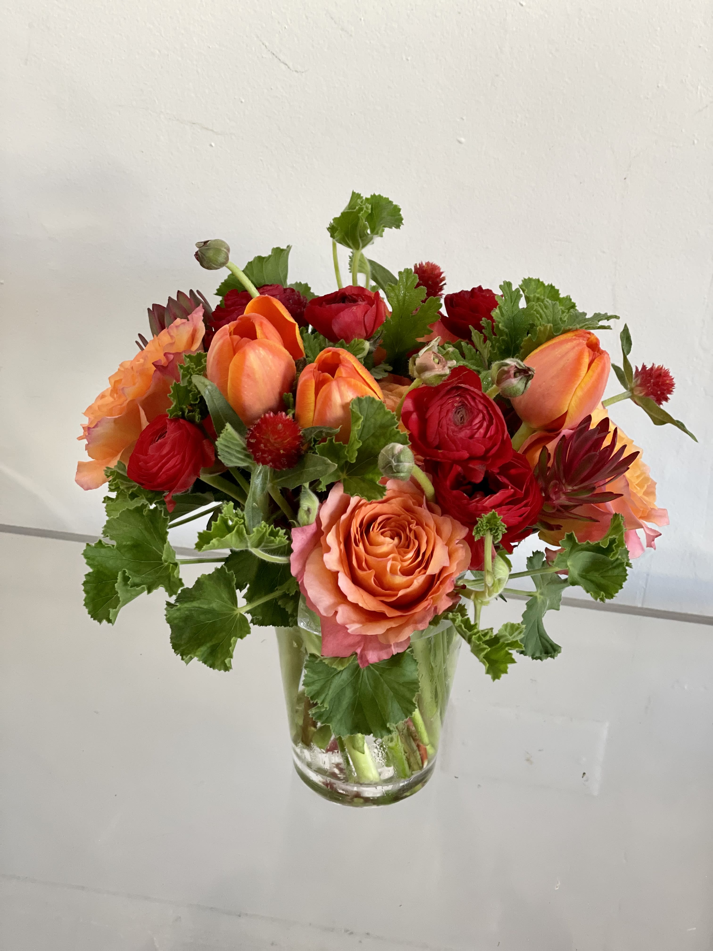 Tiny Dancer  - Designers Choice in red and orange with greens. Crisp and clean set in a glass vase These are perfect for side table, small desk, or bedside arrangement.   NOTES: We will choose the freshest and most interesting ingredients at the market to make this arrangement shine. Vessels may be substituted depending on availability.  *VALENTINE'S DAY DELIVERIES ARE LIMITED SO ORDER EARLY! PLEASE CALL THE SHOP TO PLACE ORDER IF THEY NEED TO BE TIMED* ** This is an approximate picture of what the arrangement will look like the week of Valentine's Day Weekend February 9, 2026 - February 14, 2026 ** If any design element such as color or flower/foliage are very important to your order please contact us to ensure availability first by calling the shop at 415 751 2747 