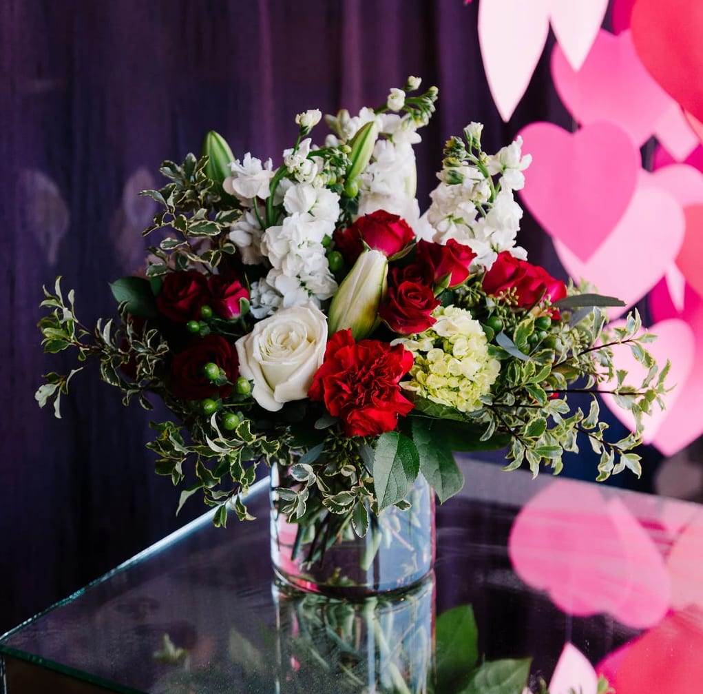 Romance in Bloom - This stunning Valentine's Day arrangement is a timeless expression of love and passion. Featuring classic red roses, symbolizing deep romance, and pure white roses for sincerity, it creates a beautiful contrast. The bouquet is enhanced with white stock flowers for elegance, vibrant red carnations for admiration, and delicate green hydrangeas adding a fresh, lush touch. Accents of greenery and unopened lilies bring texture and anticipation, while the arrangement sits in a clear glass vase for a modern yet sophisticated look.