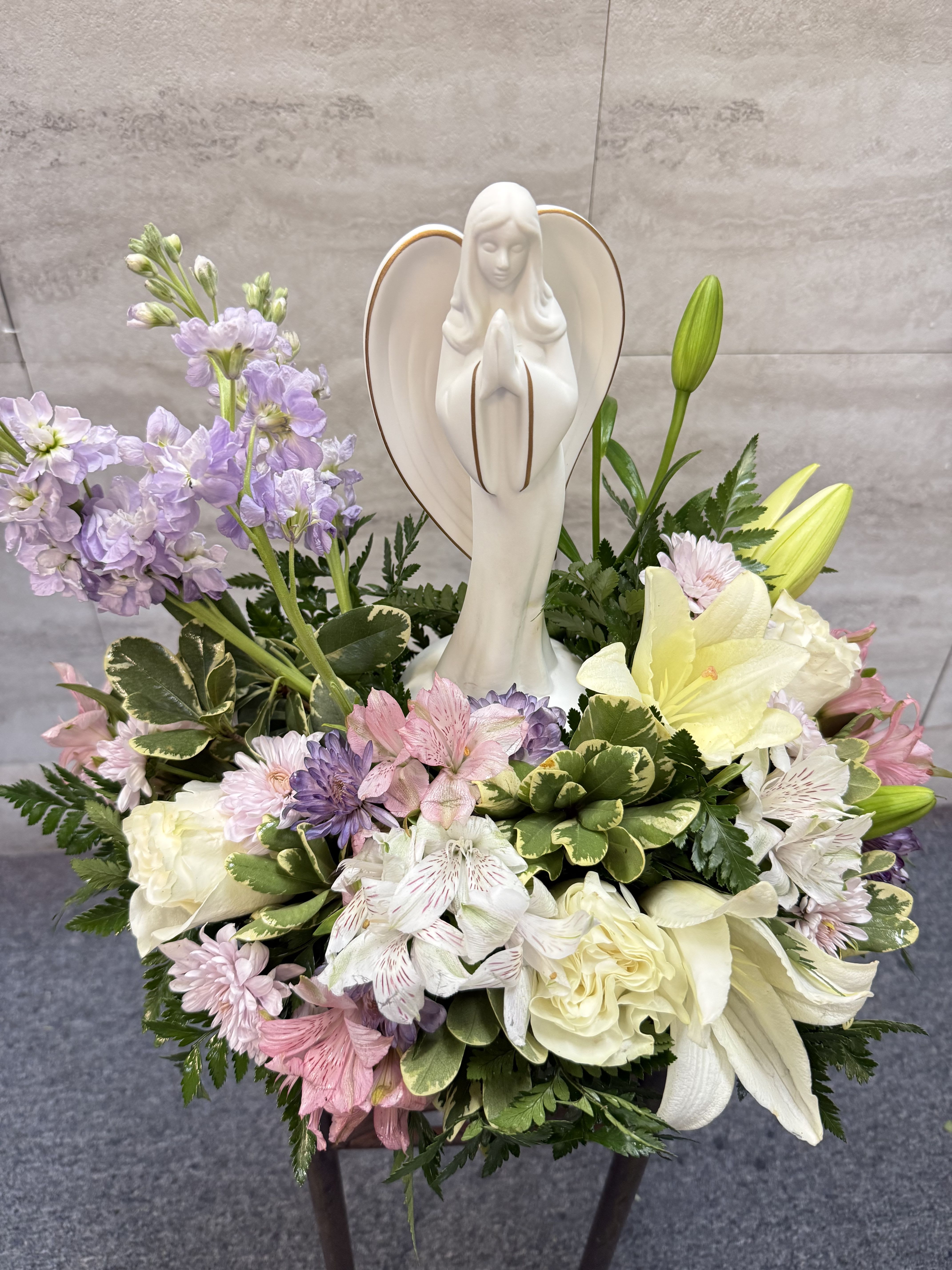 Beautiful angel  - is a serene and heartfelt arrangement featuring a soft blend of elegant blooms in gentle pastel tones. Delicate lavender stock, pink and white alstroemeria, creamy white roses, chrysanthemums, and fresh lilies are thoughtfully arranged with lush greenery to create a comforting and graceful display. At the center stands a peaceful angel figurine, symbolizing love, protection, and remembrance. **The photo shown represents the Deluxe version**, offering a fuller design with added blooms and a more abundant presentation. This arrangement provides a sense of calm and hope, making it a meaningful tribute for sympathy, memorials, or moments of reflection. 