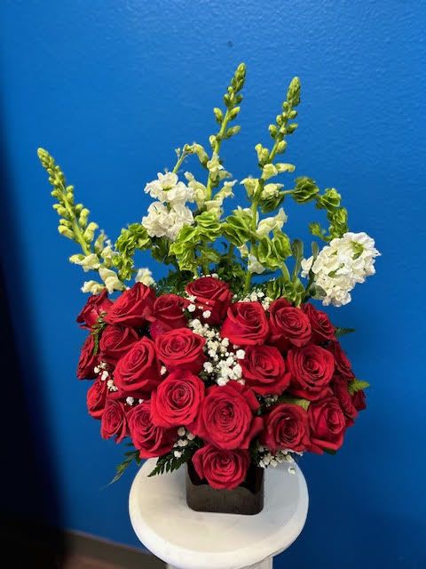 Loving emotions - A lush arrangement of deep red roses accented with white baby’s breath sits in a dark square vase on a white pedestal. Tall green stems and white snapdragons add height and elegance, giving the bouquet a full, refined look. 