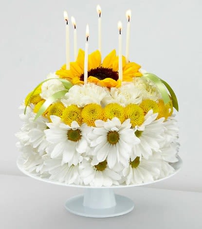 Birthday Smiles Floral Cake - Blow out the candles, and help make it their brightest birthday yet with our Birthday Smiles Floral Cake. This one–of–a–kind arrangement is frosted with a collection of bright sunflowers, yellow button pompons, white daisy pompons and white carnations. Topped with birthday candles and satin ribbon, this unique gift will bring a smile to their face and make it a truly memorable celebration.