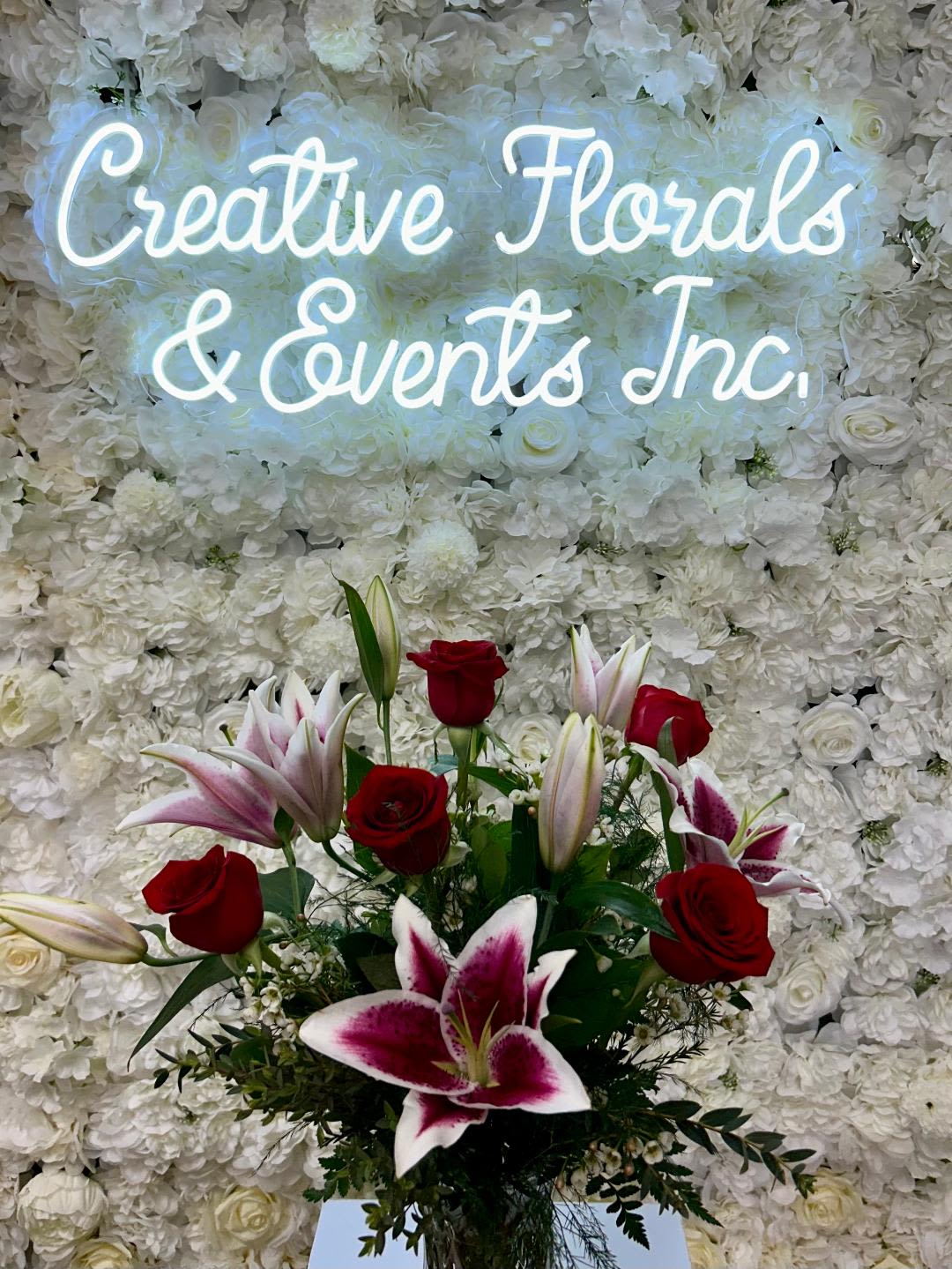 V128 - A vased arrangement of pink lilies and red roses in a tall clear glass vase with mixed greenery and filler. 