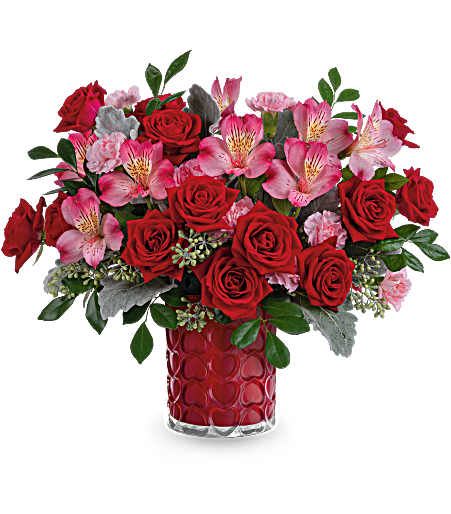 Enchanted Heart Bouquet - Adorned with a charming heart pattern and gleaming with a pearlescent finish, this delightful glass vase adds an extra touch of magic to their Valentine's Day when filled with a stunning red and pink bouquet.  This Valentine's Day arrangement features red spray roses, pink alstroemeria, and miniature light pink carnations, complemented by dusty miller, seeded eucalyptus, and huckleberry.