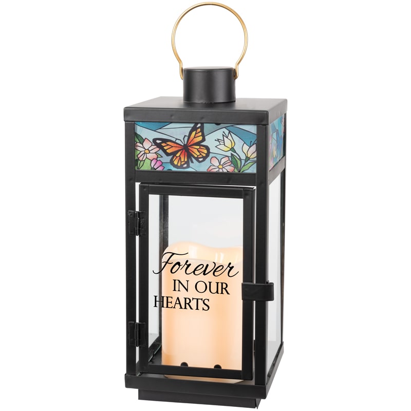 "Forever In Our Hearts" Stained Glass Top Lantern - Our stained glass lanterns are made with high-quality black finish. They come inscribed with sentimental quotes amid beautiful stained glass style artwork and a charming flameless LED candle. Suitable for hanging or use as a freestanding item.  They are weather friendly! Butterfly on the stain with blue around the glass brown and black butterfly and little pick flowers  Lantern Reads: "Forever In Our Heart W: 4.25" H: 12" D: 4.25"  