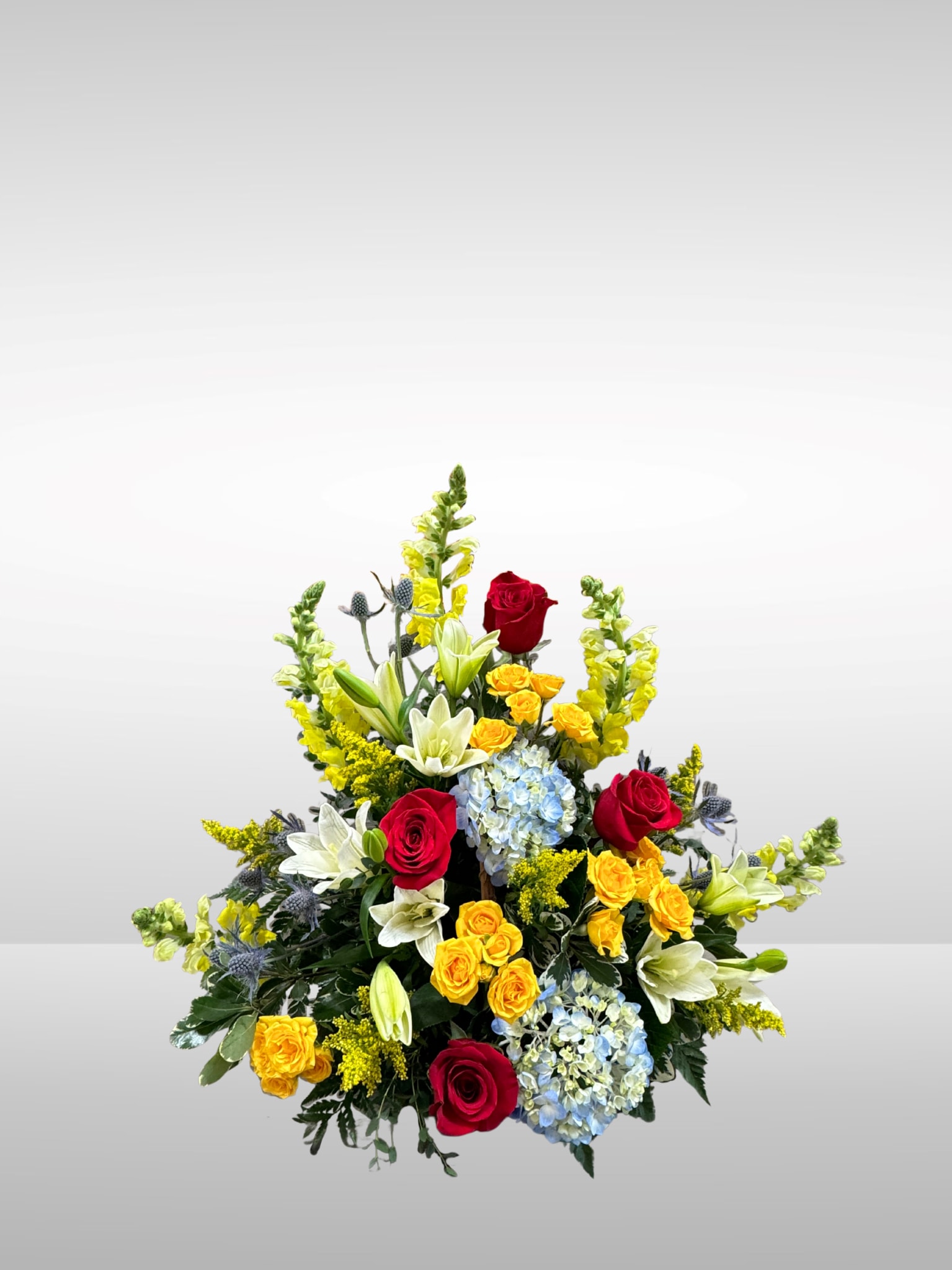 Graceful Blooms Basket - Bursting with vibrant reds, sunny yellows, whites, and soft blues, this cheerful basket of fresh blooms offers both comfort and joy. Whether shared in remembrance, given as a thoughtful gift to a friend or coworker, or simply used to brighten a space, this all-around floral arrangement brings beauty and warmth to any occasion. Set in a classic basket, it’s a heartfelt gesture that also makes a stunning centerpiece. . White lilly's, red roses with yellow spray roses, statice, blue hydrangeas, yellow snapdragons, and thistle are threaded through arranged in a wicker basket.