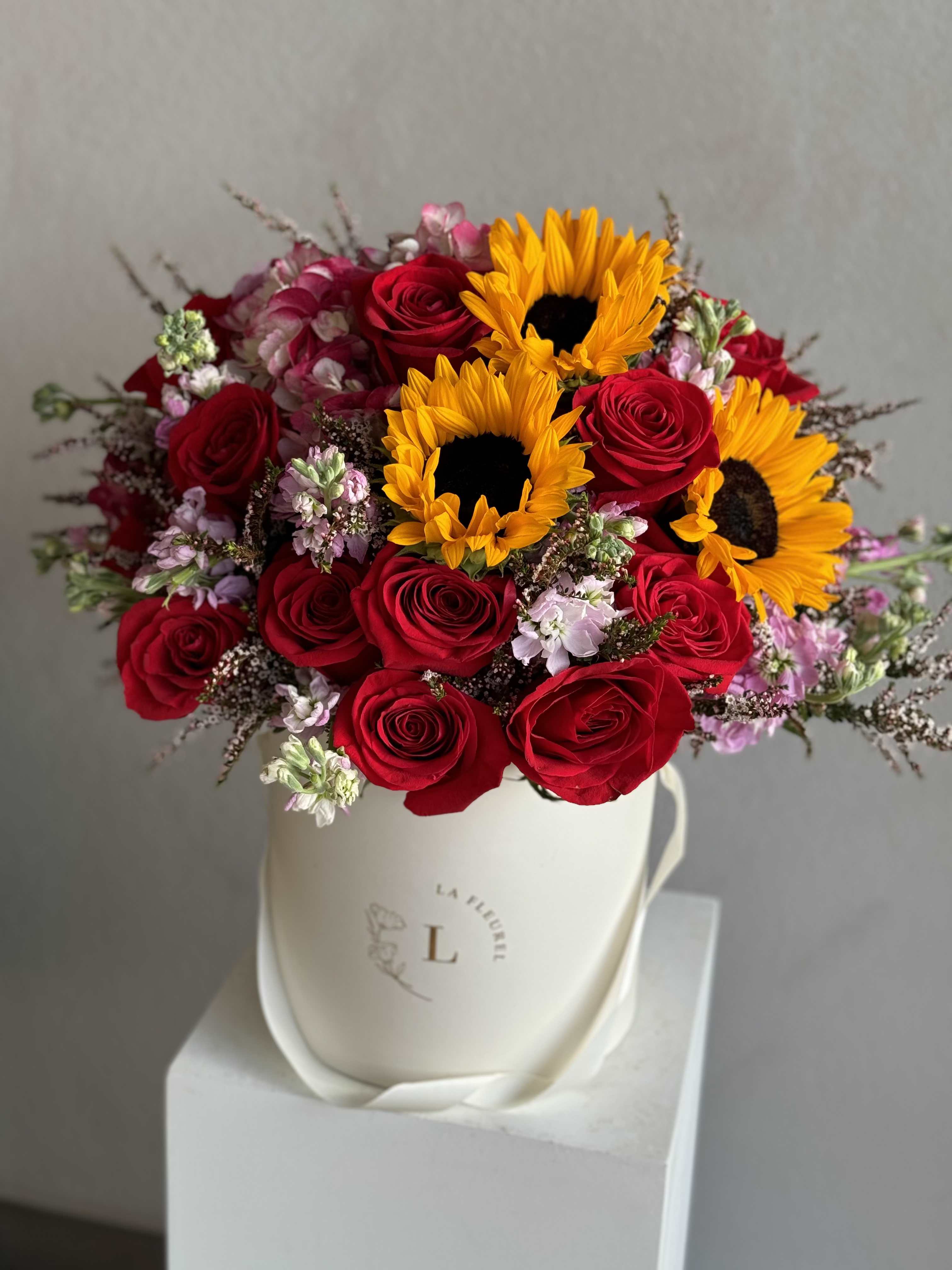 Sunset Romance - A captivating arrangement that combines the rich, romantic allure of red roses with the cheerful brightness of sunflowers, complemented by a mix of complementary blooms. All beautifully arranged in a pristine white box, this bouquet captures the essence of a sunset’s warm embrace, bringing a touch of elegance and joy to any occasion.